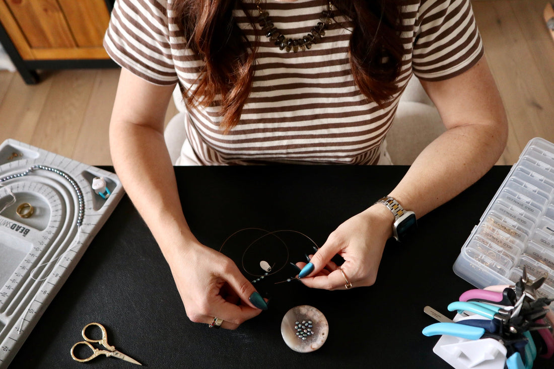 Auréad Designs founder stringing a handmade gemstone necklace at her jewelry-making workspace with beading board, pliers, and gemstone beads