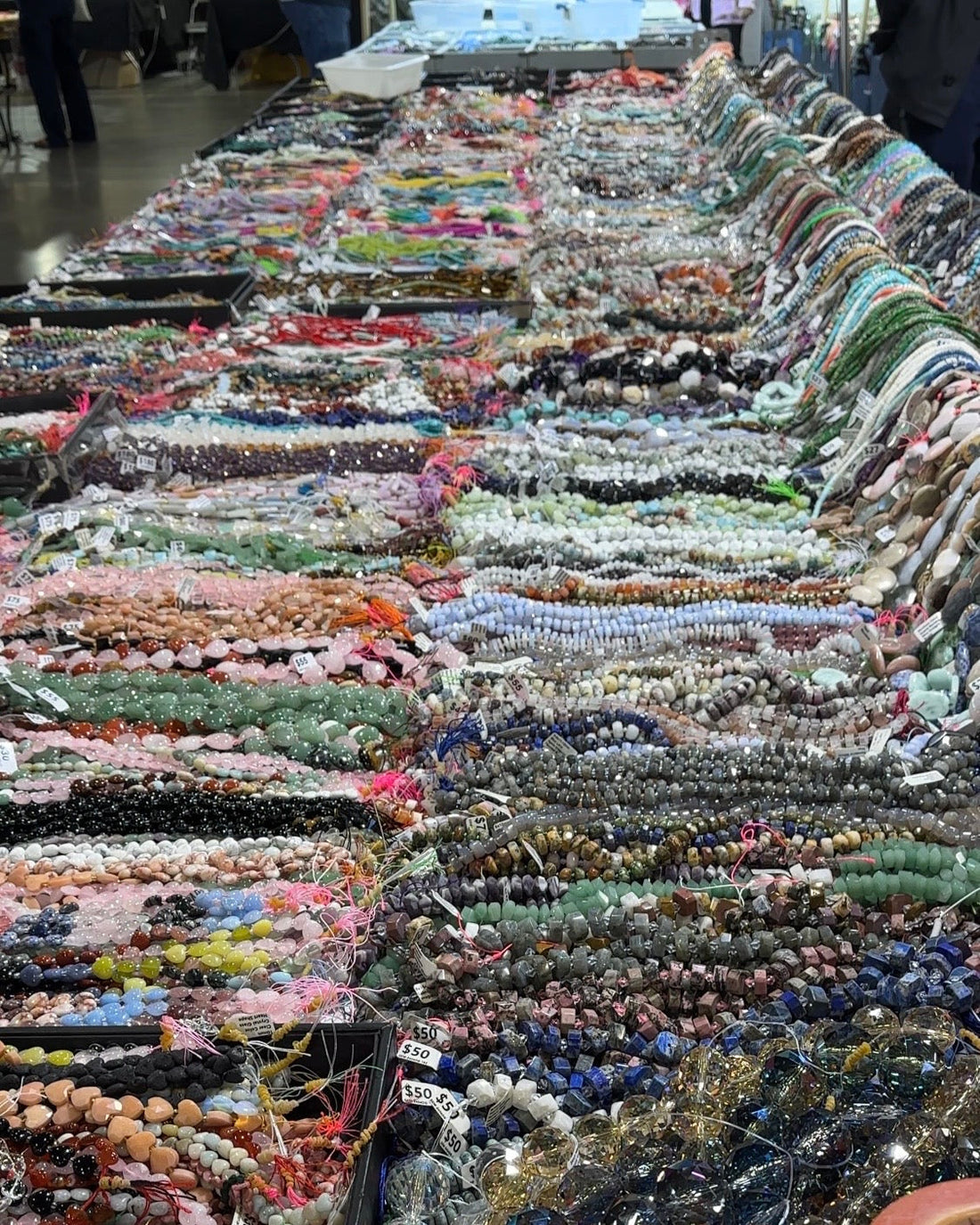 Long gem faire vendor table covered in rows of colorful gemstone bead strands, the sourcing starting point for Auréad Designs handmade jewelry