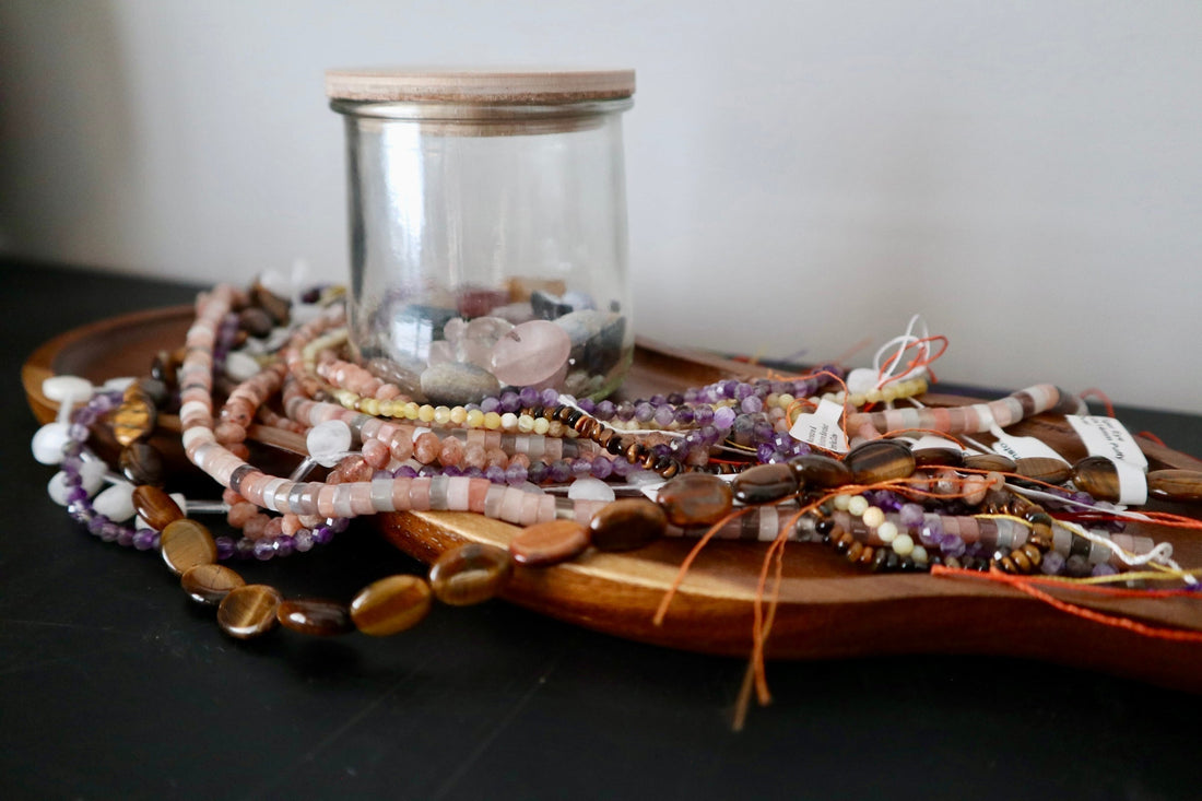 Gemstone bead strands including amethyst, tiger eye, and opal beads arranged on a round wood tray with a glass jar of loose gemstone chips, Auréad Designs sourcing haul