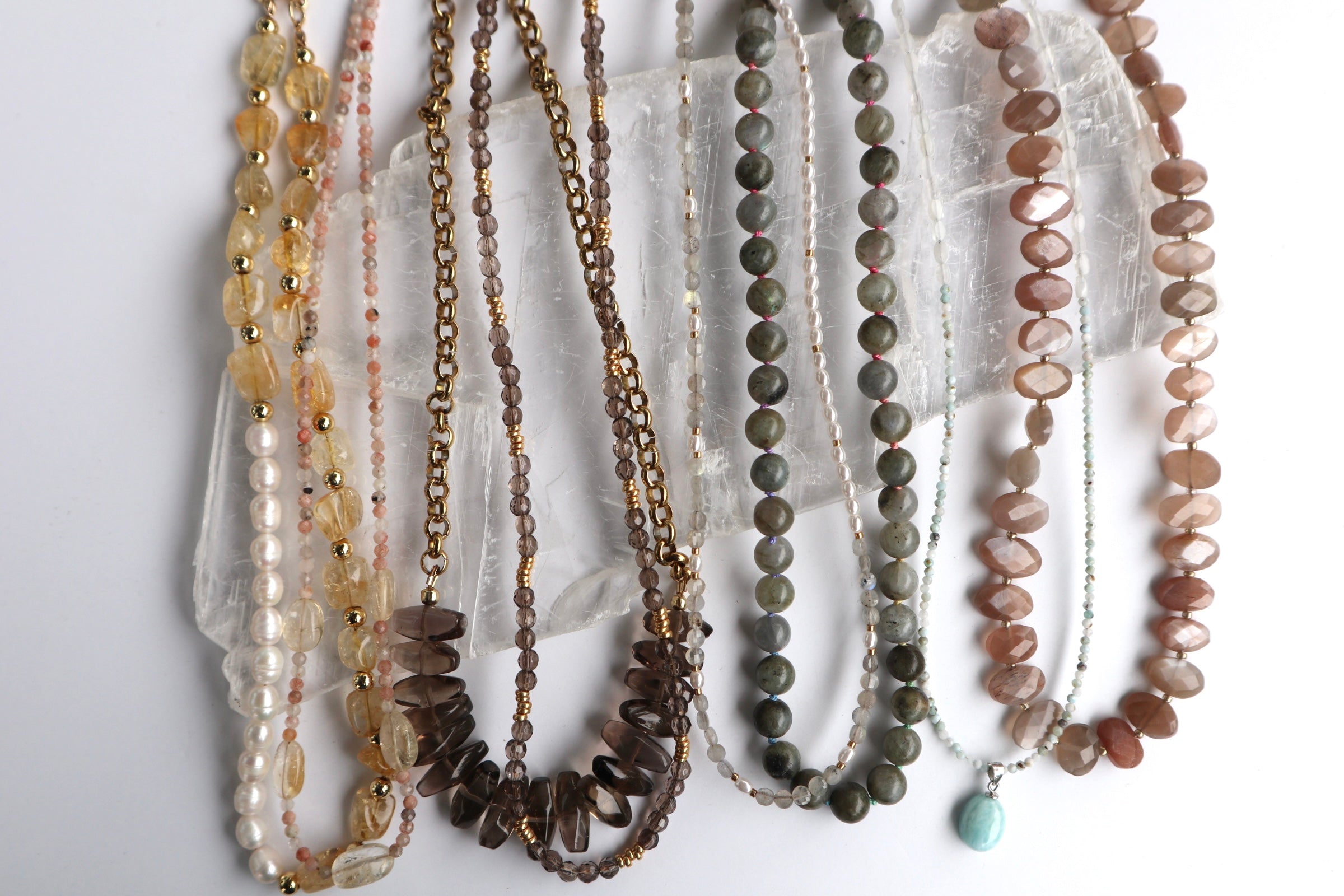 Editorial flat lay of seven Auréád Designs gemstone necklaces arranged across a white selenite slab, featuring citrine, sunstone, smoky quartz, labradorite, moonstone, and peach moonstone in warm earth tones