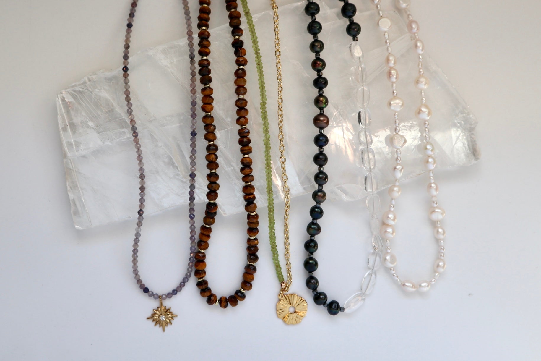 Handmade gemstone necklaces by Auréad Designs laid on a white selenite slab including Astrielle iolite star pendant, Sable tiger eye, Florae peridot gold chain, Tashira clear quartz and black pearl, and Aveline freshwater pearl necklaces