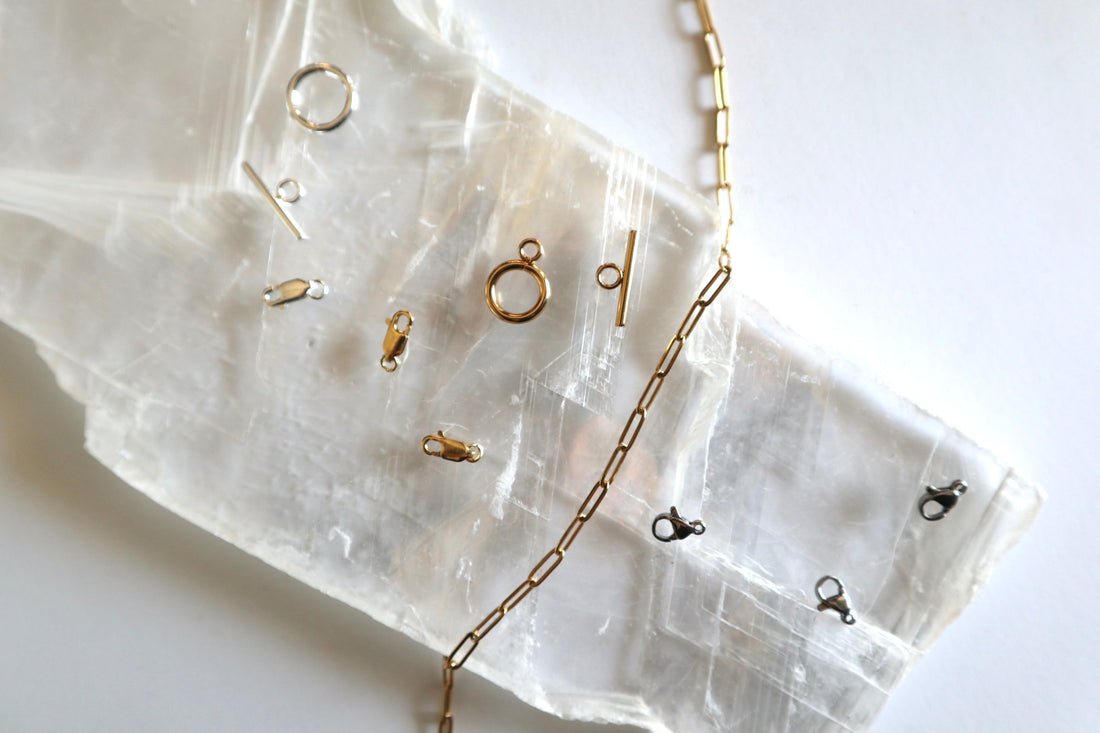 Flat lay of jewelry hardware including lobster clasps, toggle clasps, and gold paperclip chain on white selenite slab, Auréad Designs