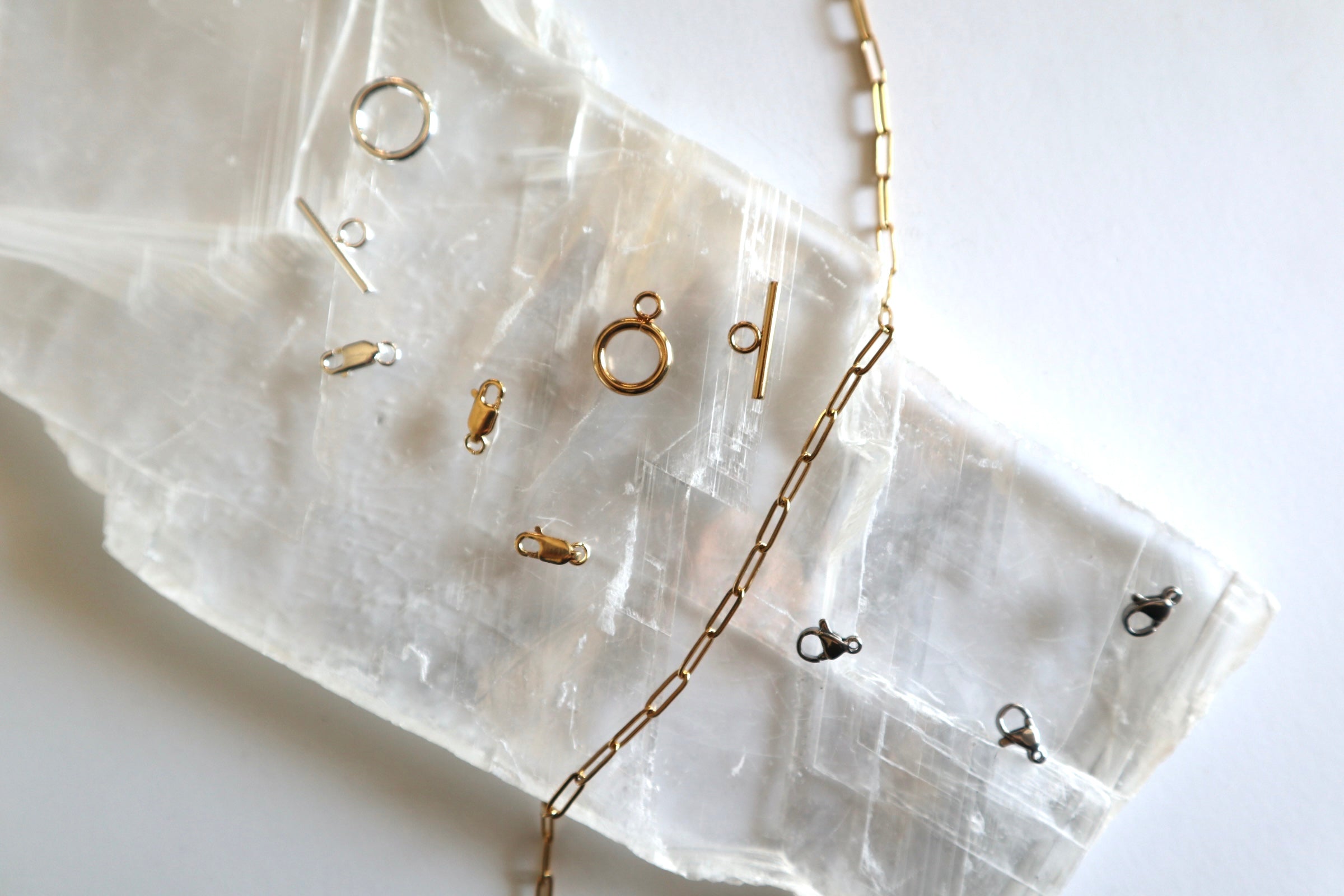 Flat lay of jewelry hardware including lobster clasps, toggle clasps, and gold paperclip chain on white selenite slab, Auréad Designs