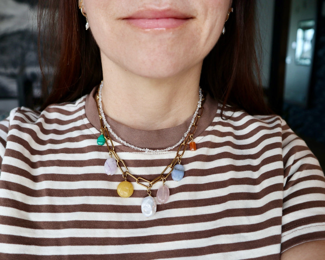 Layered handmade gemstone necklaces worn over a striped tee, including a gold paperclip chain with colorful gemstone charms and a delicate crystal beaded necklace by Auréád Designs