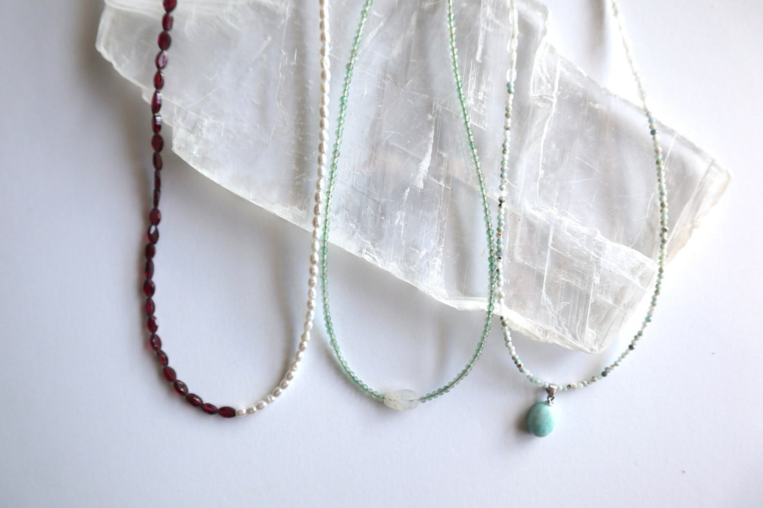 Flat lay of three Auréad Designs handmade gemstone necklaces on a white selenite slab — Garnelle in faceted garnet, Aveline in freshwater pearl, and Primelle in green aventurine with a moonstone focal, styled as meaningful graduation gift ideas.