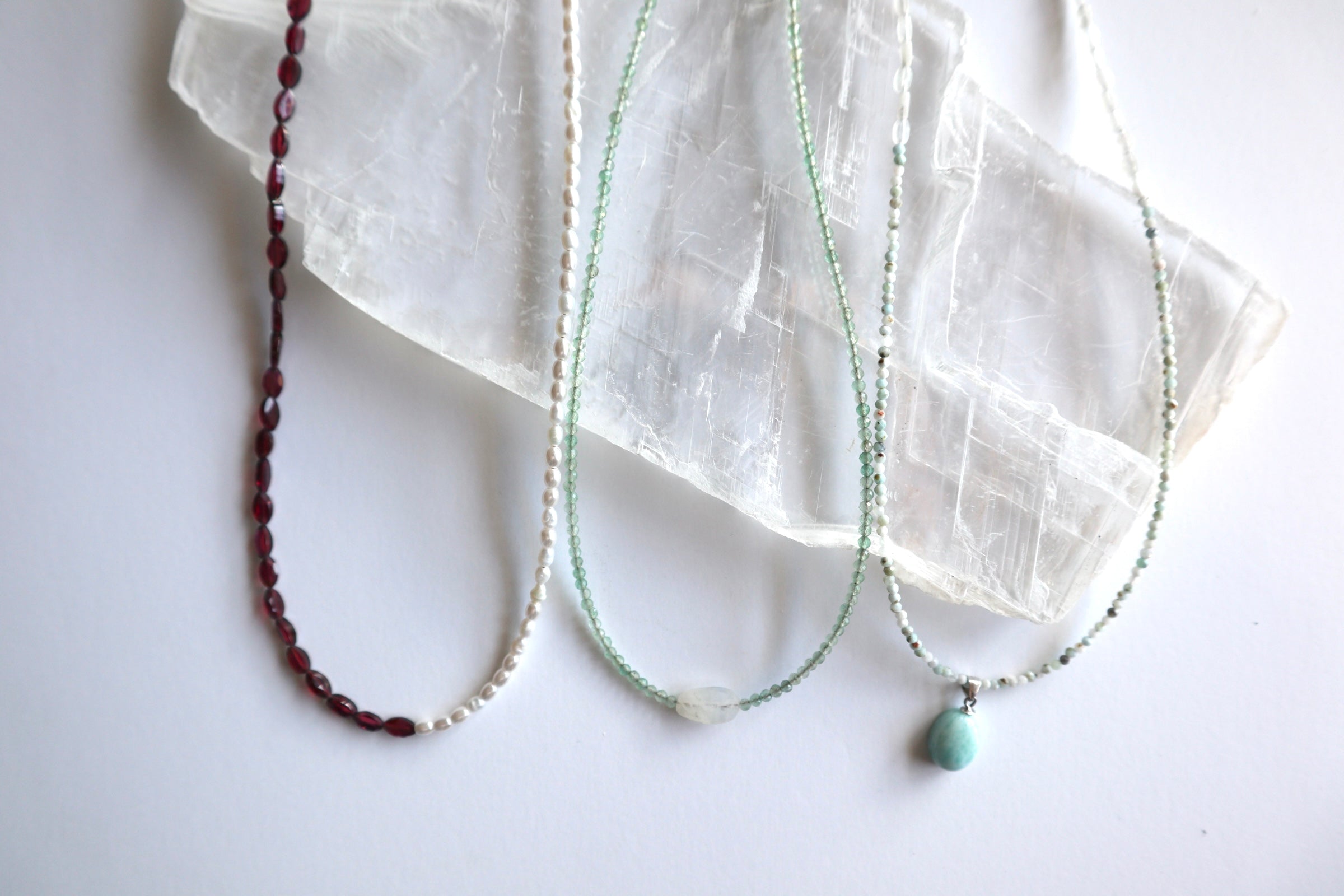 Flat lay of three Auréad Designs handmade gemstone necklaces on a white selenite slab — Garnelle in faceted garnet, Aveline in freshwater pearl, and Primelle in green aventurine with a moonstone focal, styled as meaningful graduation gift ideas.
