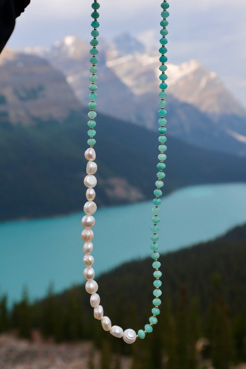 Lake necklace by Auréad Designs held against Peyto Lake in Banff National Park, showing the asymmetric amazonite rondelle and coin freshwater pearl strand with the turquoise glacially-fed water behind it, Glacier Collection