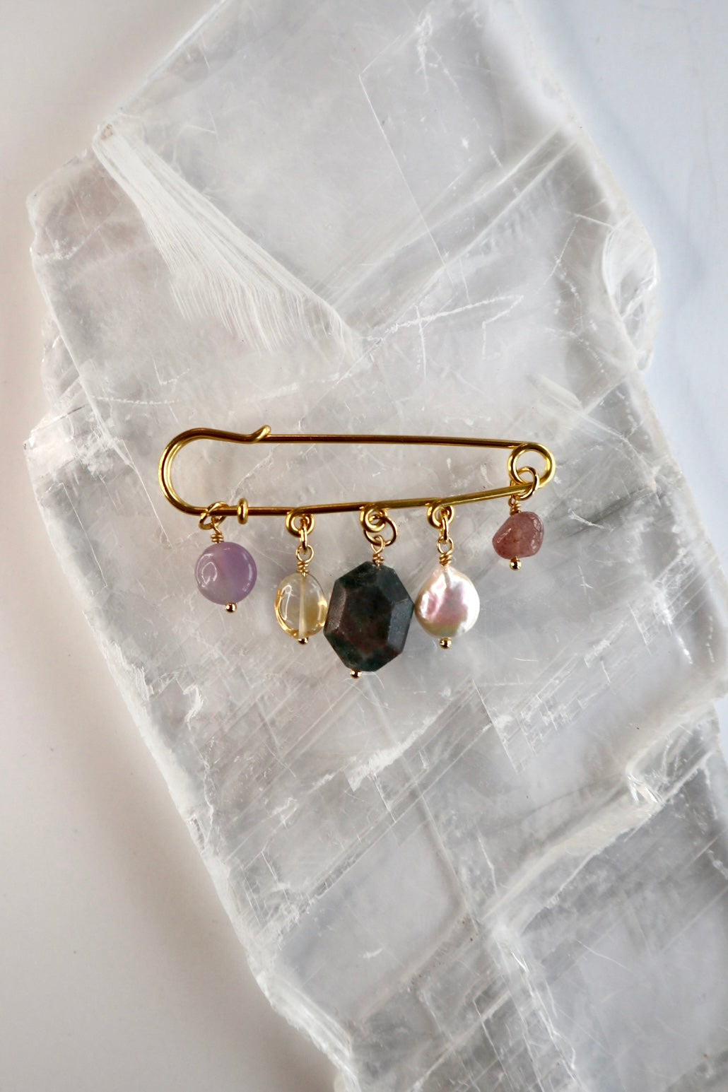 Amorine mixed gemstone brooch by Auréád Designs resting on a white selenite slab, featuring five charm drops of amethyst, citrine, apatite, freshwater pearl, and strawberry quartz hanging from a gold stainless steel pin