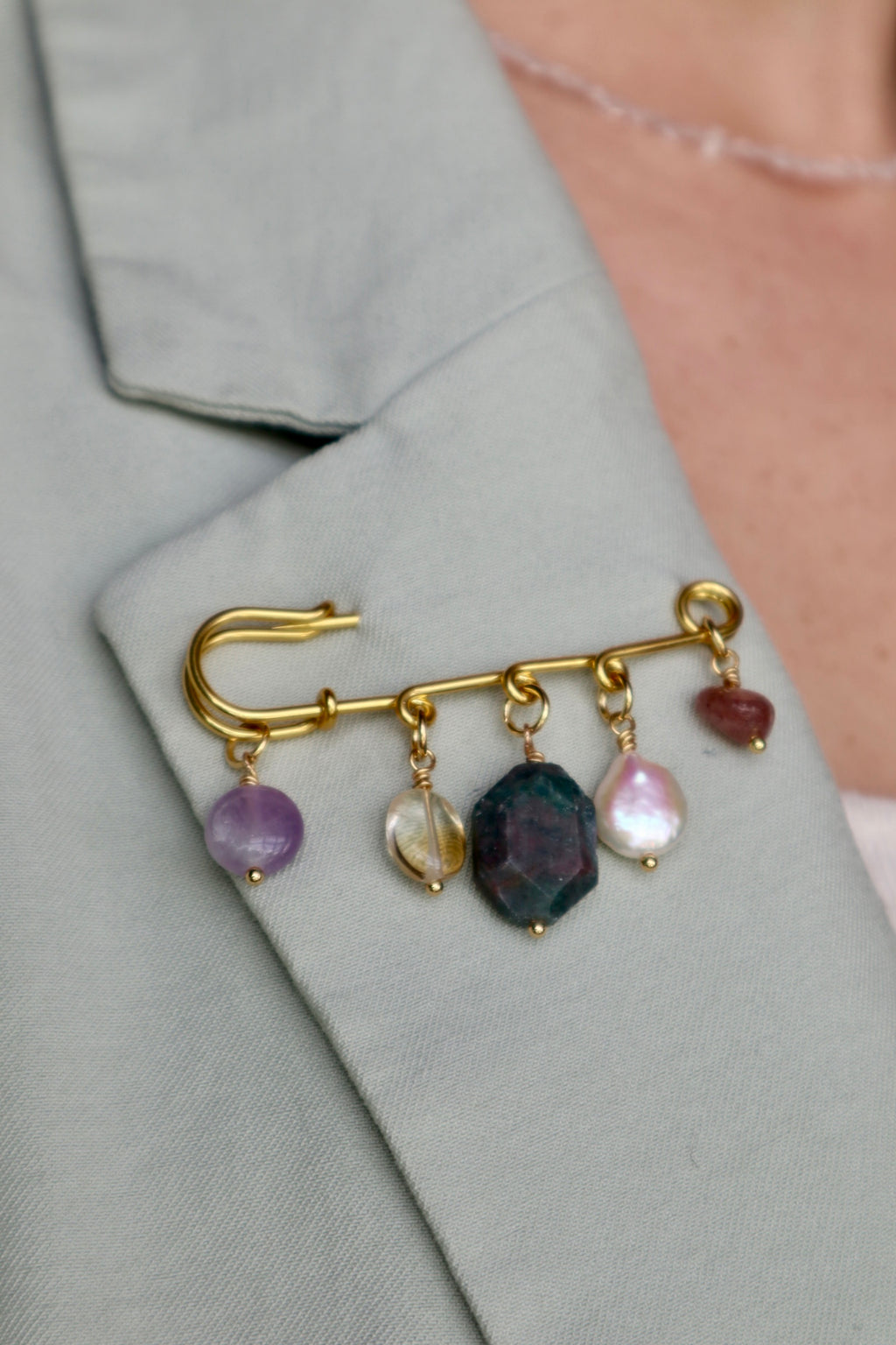Amorine mixed gemstone brooch by Auréád Designs pinned to the lapel of a sage green blazer, showing amethyst, citrine, apatite, freshwater pearl, and strawberry quartz charm drops against the fabric