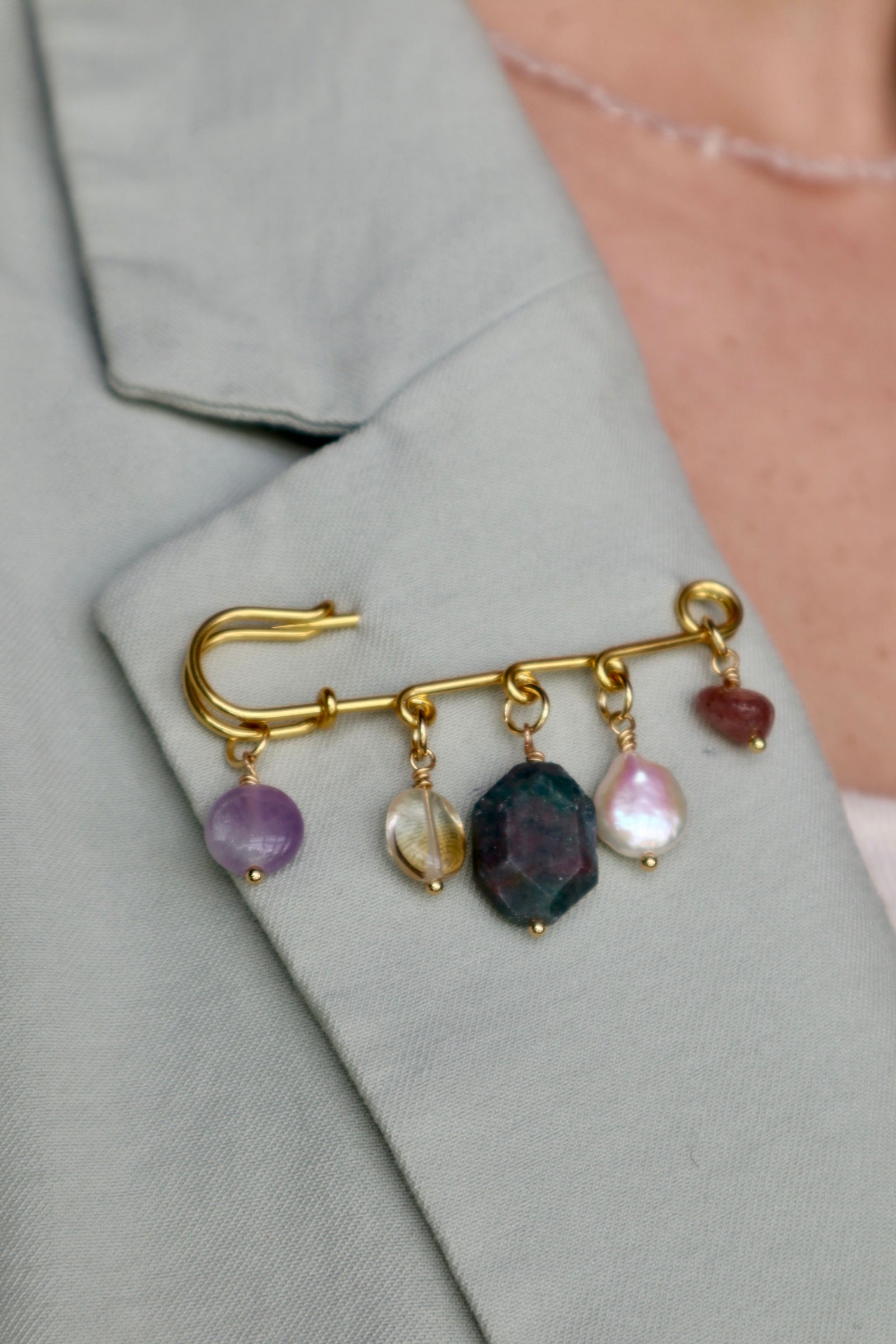 Amorine mixed gemstone brooch by Auréád Designs pinned to the lapel of a sage green blazer, showing amethyst, citrine, apatite, freshwater pearl, and strawberry quartz charm drops against the fabric
