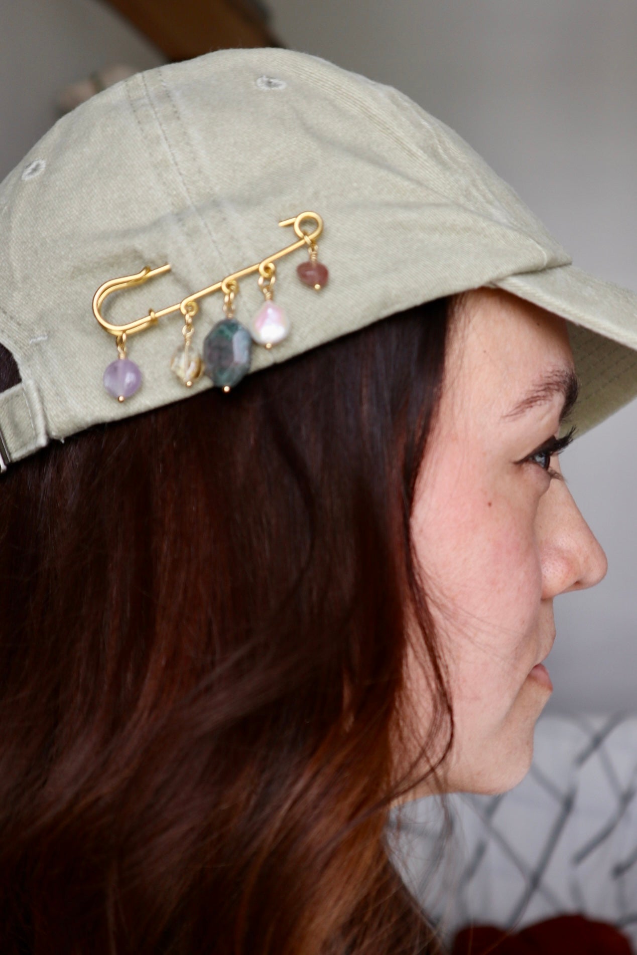 Amorine mixed gemstone brooch by Auréád Designs pinned to the brim of a sage green baseball cap, showing amethyst, citrine, apatite, freshwater pearl, and strawberry quartz charm drops
