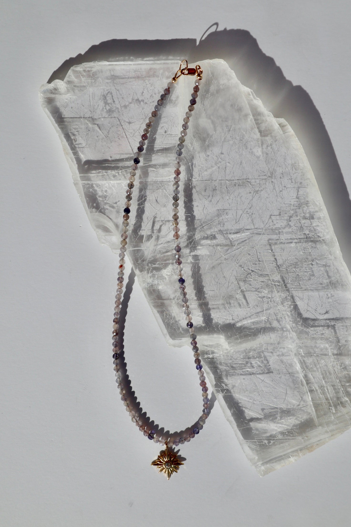 Astrielle necklace by Auréad Designs, featuring micro-faceted iolite gemstone beads and a gold radiant star pendant with a 14k gold-filled lobster clasp, laid flat on a white selenite slab
