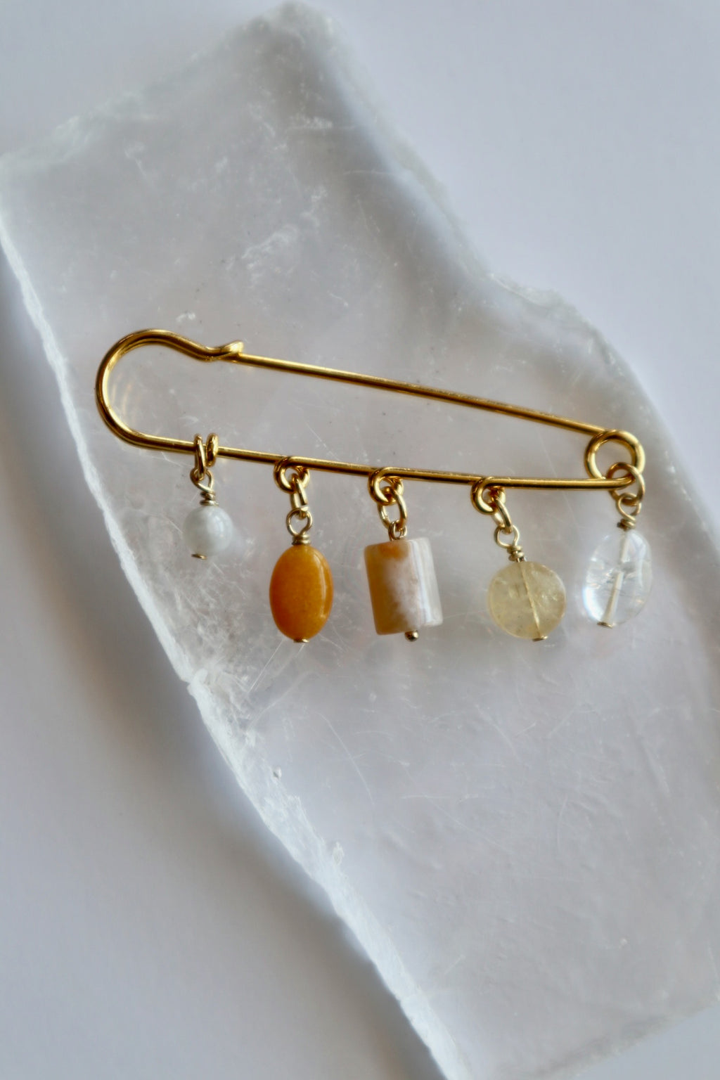 Auréad Designs Aurene gold kilt pin brooch with rainbow moonstone, yellow jade, cherry blossom agate, citrine, and clear quartz charm drops, overhead flat lay on white selenite slab.