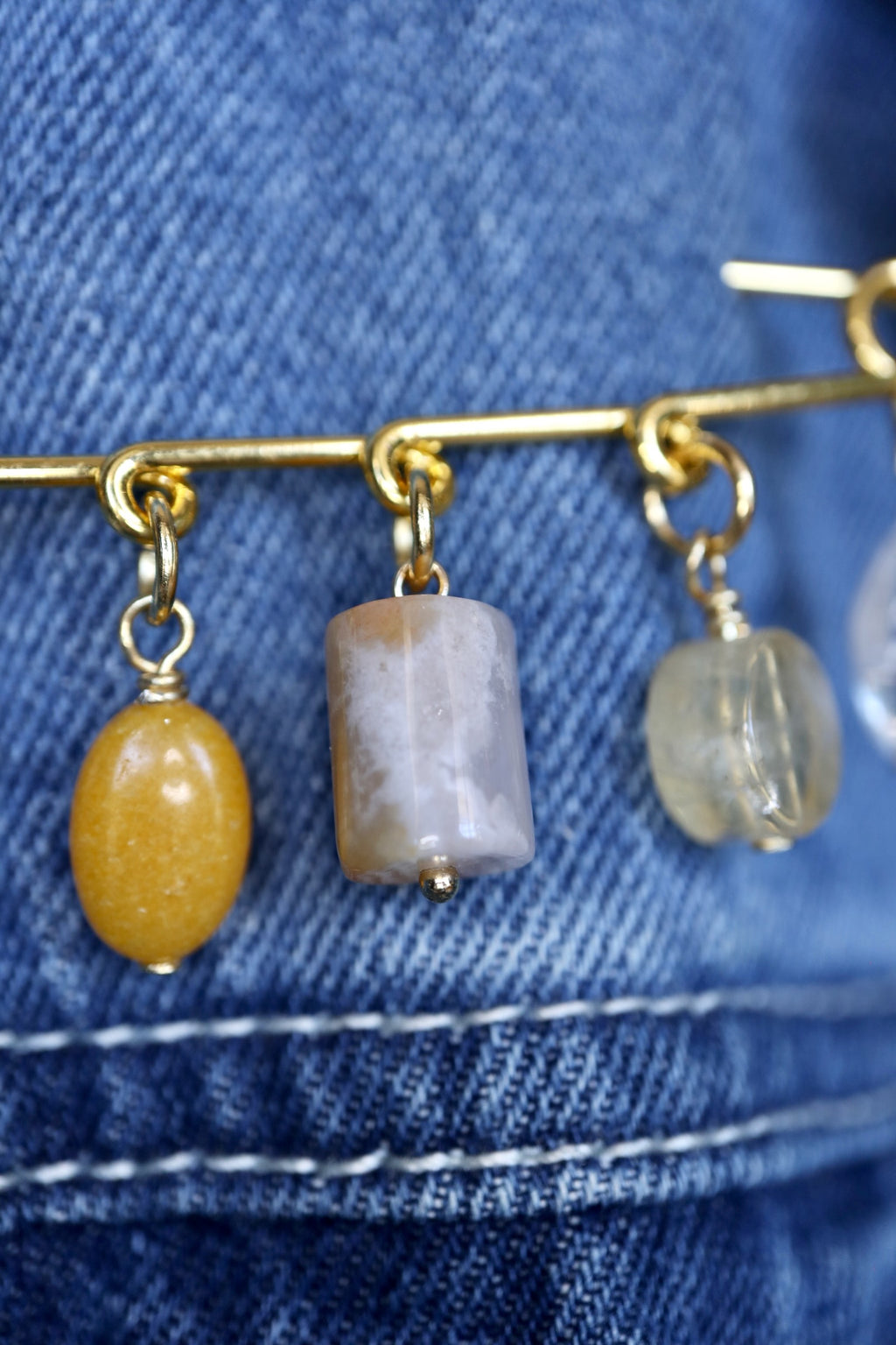 Macro detail of Auréad Designs Aurene gold kilt pin brooch showing yellow jade oval and cherry blossom agate cylinder charm drops with gold jump ring hardware, worn on denim.