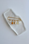 Auréad Designs Aurene gold kilt pin brooch with five dangling gemstone charms in rainbow moonstone, yellow jade, cherry blossom agate, citrine, and clear quartz, flat lay on white selenite slab.