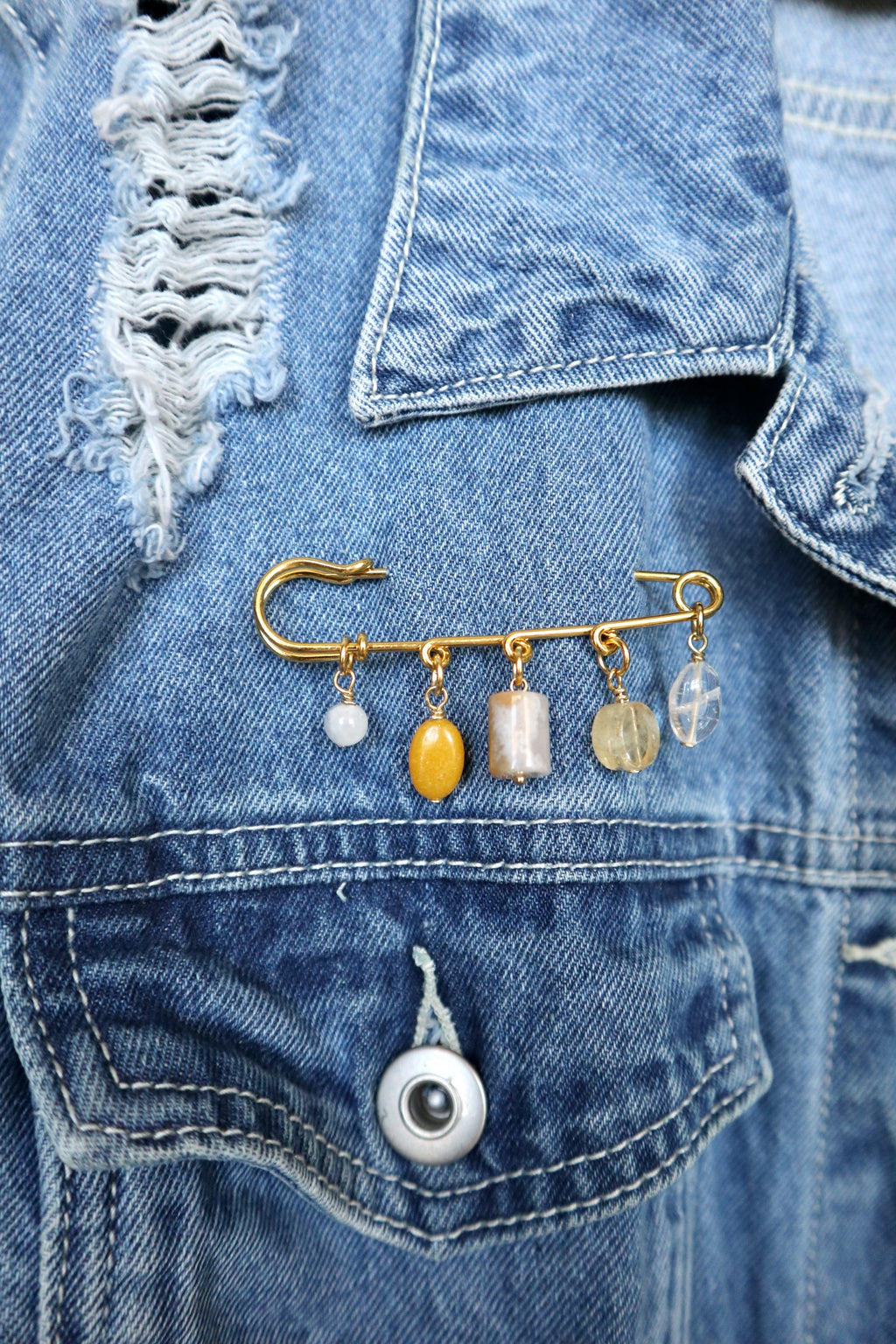 Auréad Designs Aurene gold kilt pin brooch with five gemstone charms in rainbow moonstone, yellow jade, cherry blossom agate, citrine, and clear quartz, worn on a distressed blue denim jacket.