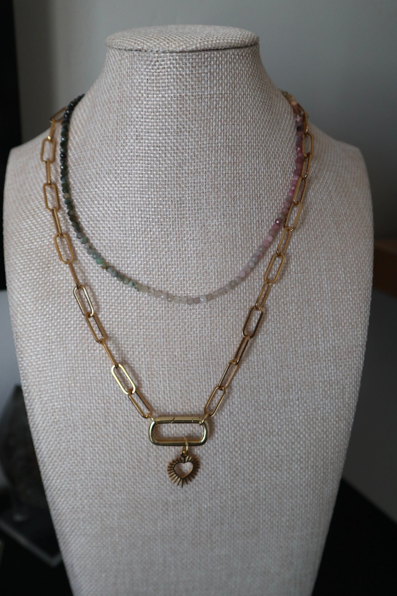 Aurora tourmaline gradient necklace by Auréad Designs displayed on a linen bust form, layered with a gold paperclip chain necklace with a heart pendant, showing the full color spectrum of multicolored micro-faceted tourmaline beads