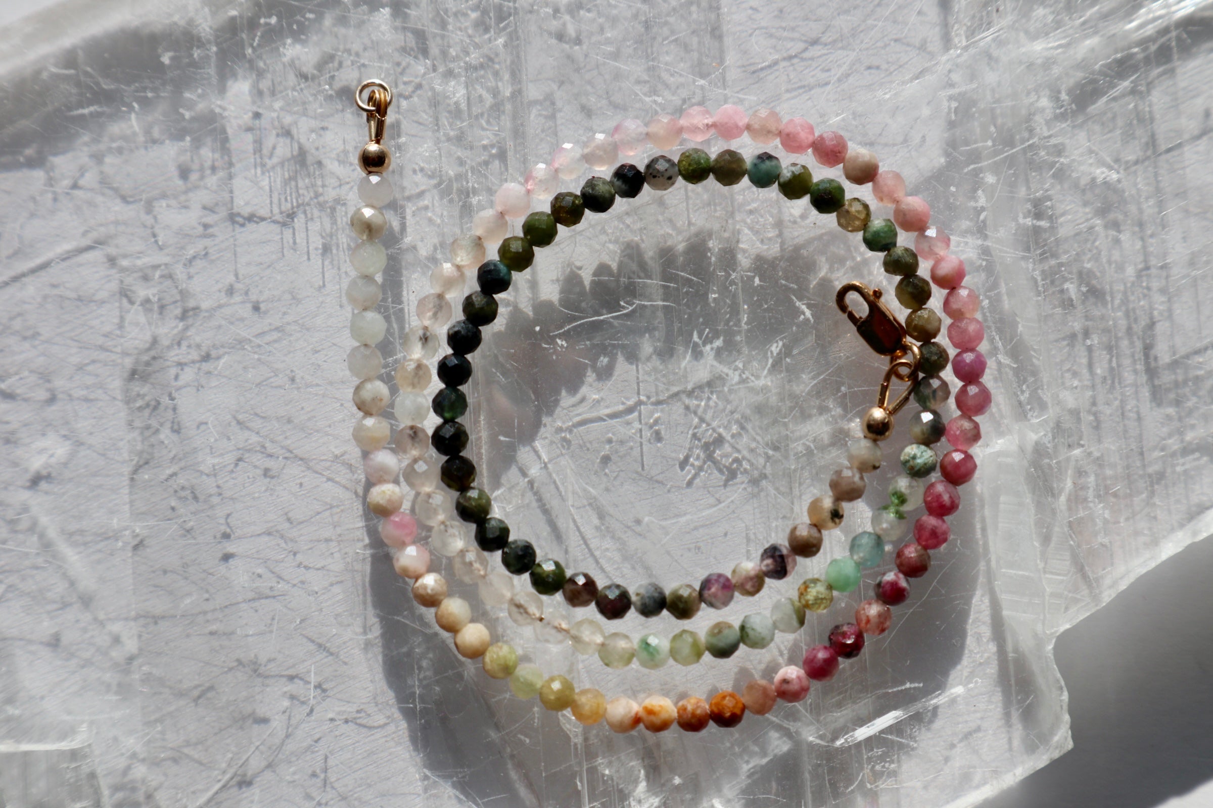 Aurora necklace by Auréad Designs coiled on a white selenite slab, displaying the full multicolored tourmaline gradient from deep green through pink, peach, and coral tones