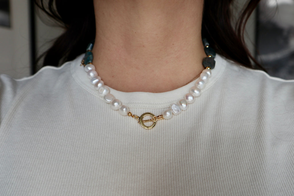 Azurea necklace by Auréad Designs worn at the collarbone, featuring freshwater pearls and a gold toggle clasp front-facing with teal gemstone beads visible at the shoulders, styled over a white top