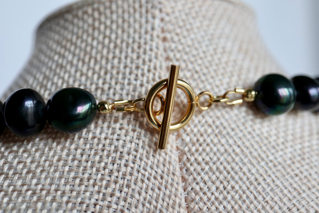 Close-up of the Azurea Noir necklace on a linen bust form, showing the gold stainless steel toggle clasp worn at the front as a focal point with deep iridescent black pearls on either side
