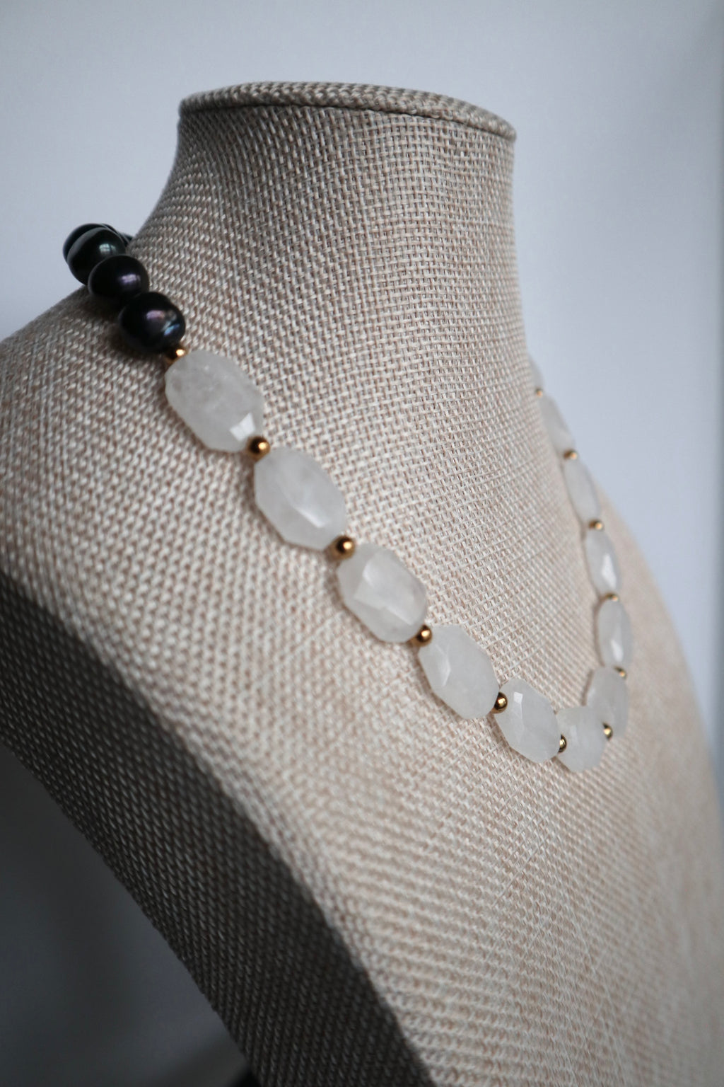 Azurea Noir necklace by Auréad Designs displayed at an angle on a linen bust form, showing the full drape of large clear quartz beads with gold spacers contrasting against the cluster of black pearls