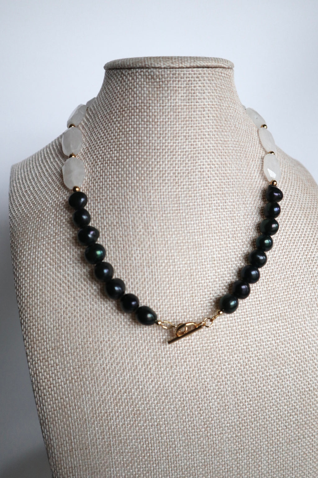 Azurea Noir necklace by Auréad Designs displayed on a linen bust form, showing the asymmetrical design with large clear quartz beads across the top and black pearls draping below, with the gold toggle clasp worn at the front
