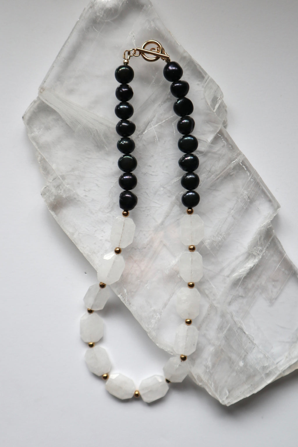 Azurea Noir necklace by Auréad Designs, featuring clear quartz beads and black pearls with gold stainless steel spacer beads and a toggle clasp, laid flat on a white selenite slab