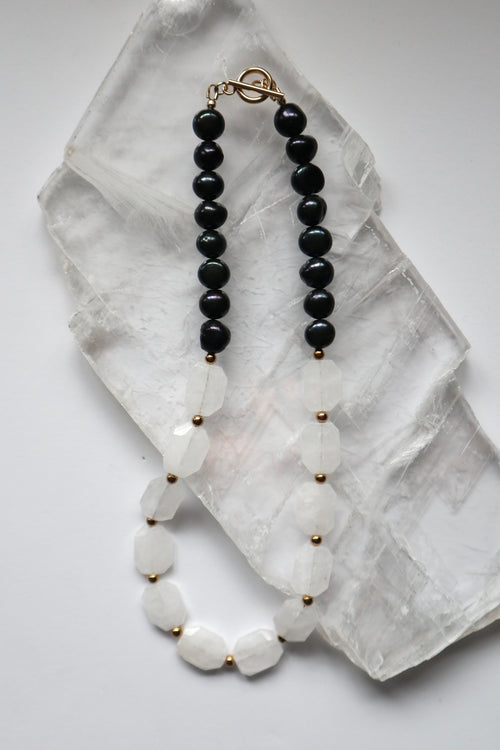 Azurea Noir necklace by Auréad Designs, featuring clear quartz beads and black pearls with gold stainless steel spacer beads and a toggle clasp, laid flat on a white selenite slab