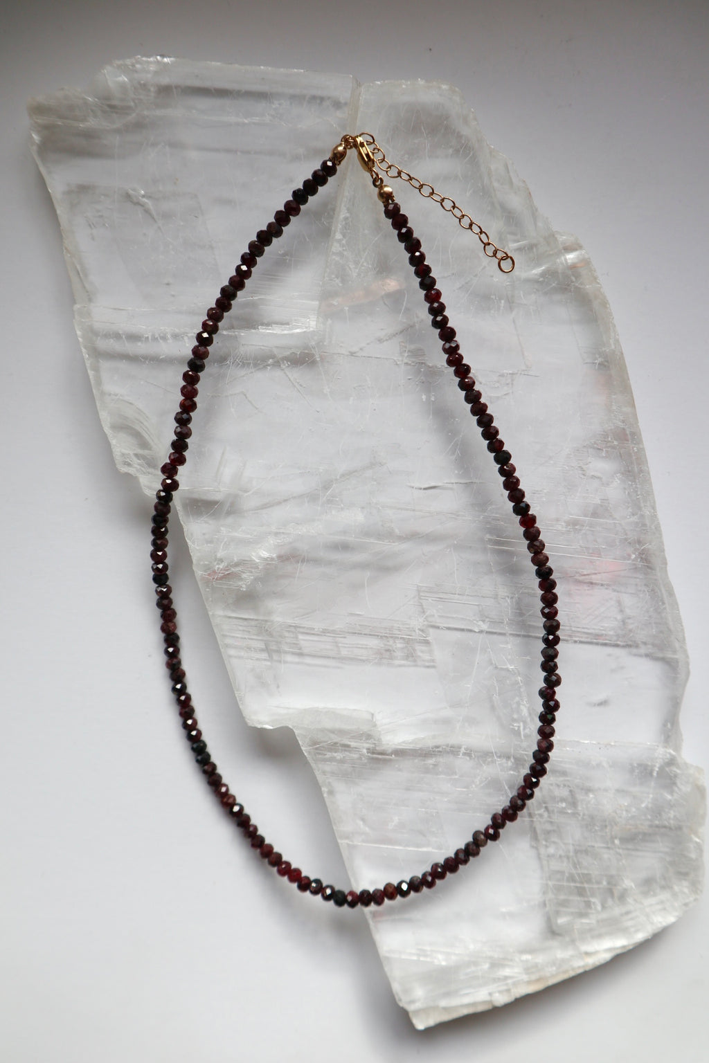 Brenna necklace by Auréad Designs, featuring faceted garnet gemstone beads with a 14k gold-filled lobster clasp and two-inch extender chain, laid flat on a white selenite slab