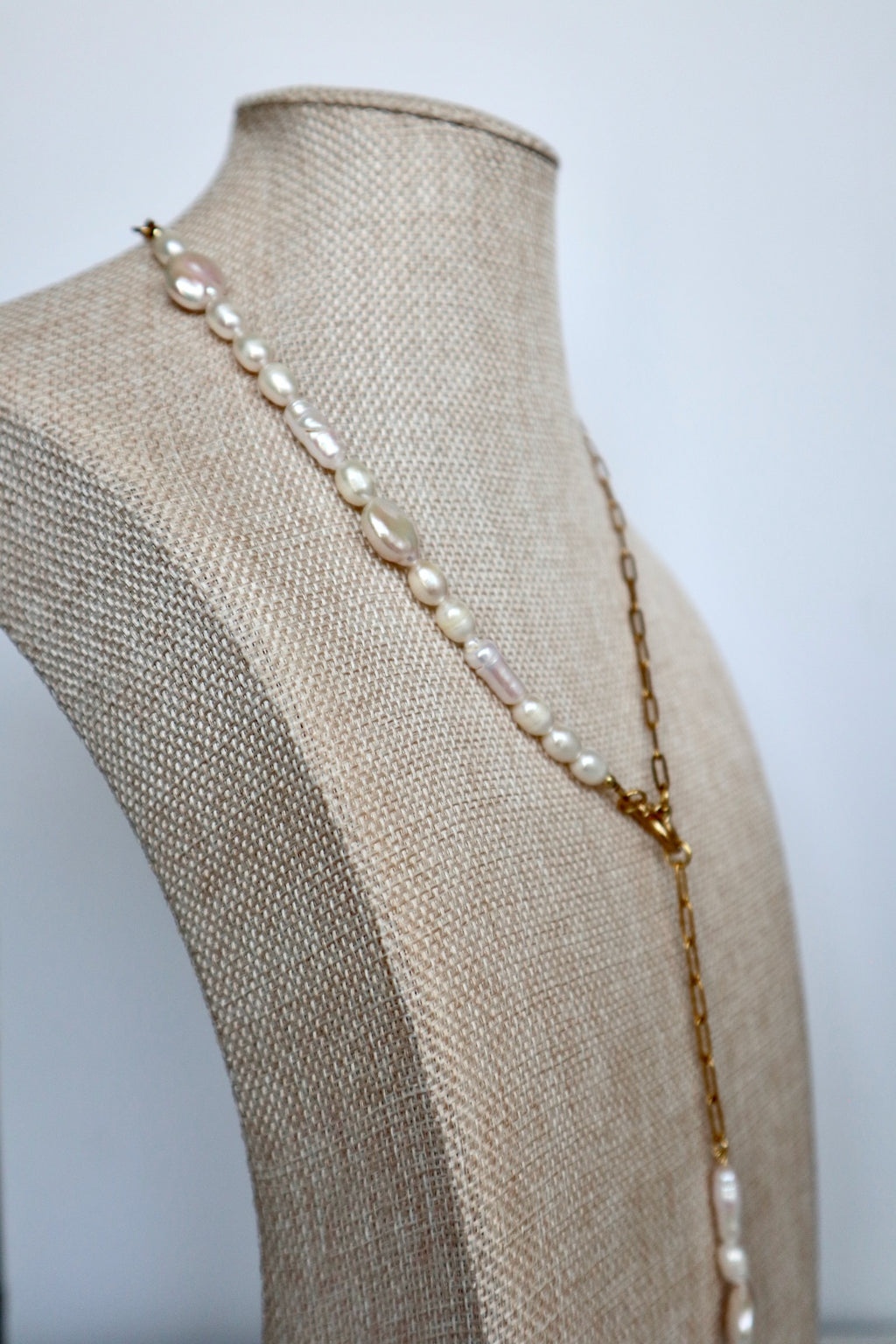 Auréad Designs Caelova lariat necklace draped at an angle on linen bust form, showing the contrast between the freshwater pearl strand half and gold paperclip chain half with pendant drop
