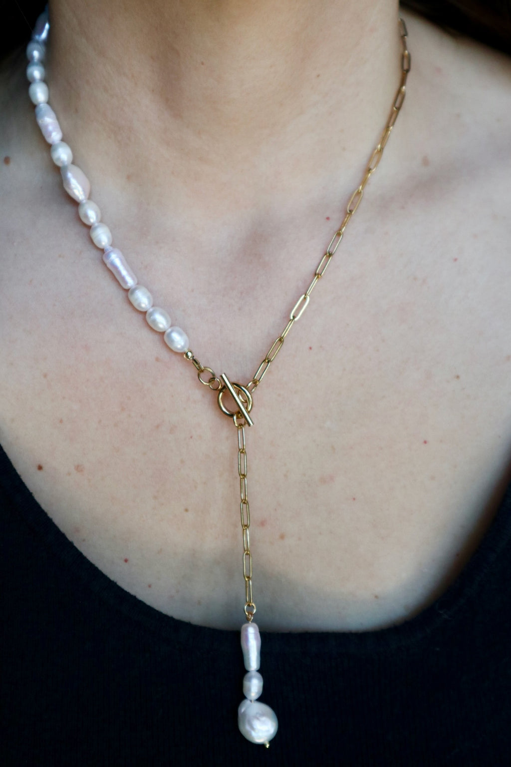 Auréad Designs Caelova lariat necklace worn on a black ribbed top, showing freshwater pearl strand on left, gold paperclip chain on right, toggle clasp at center front, and triple freshwater pearl pendant dropping below