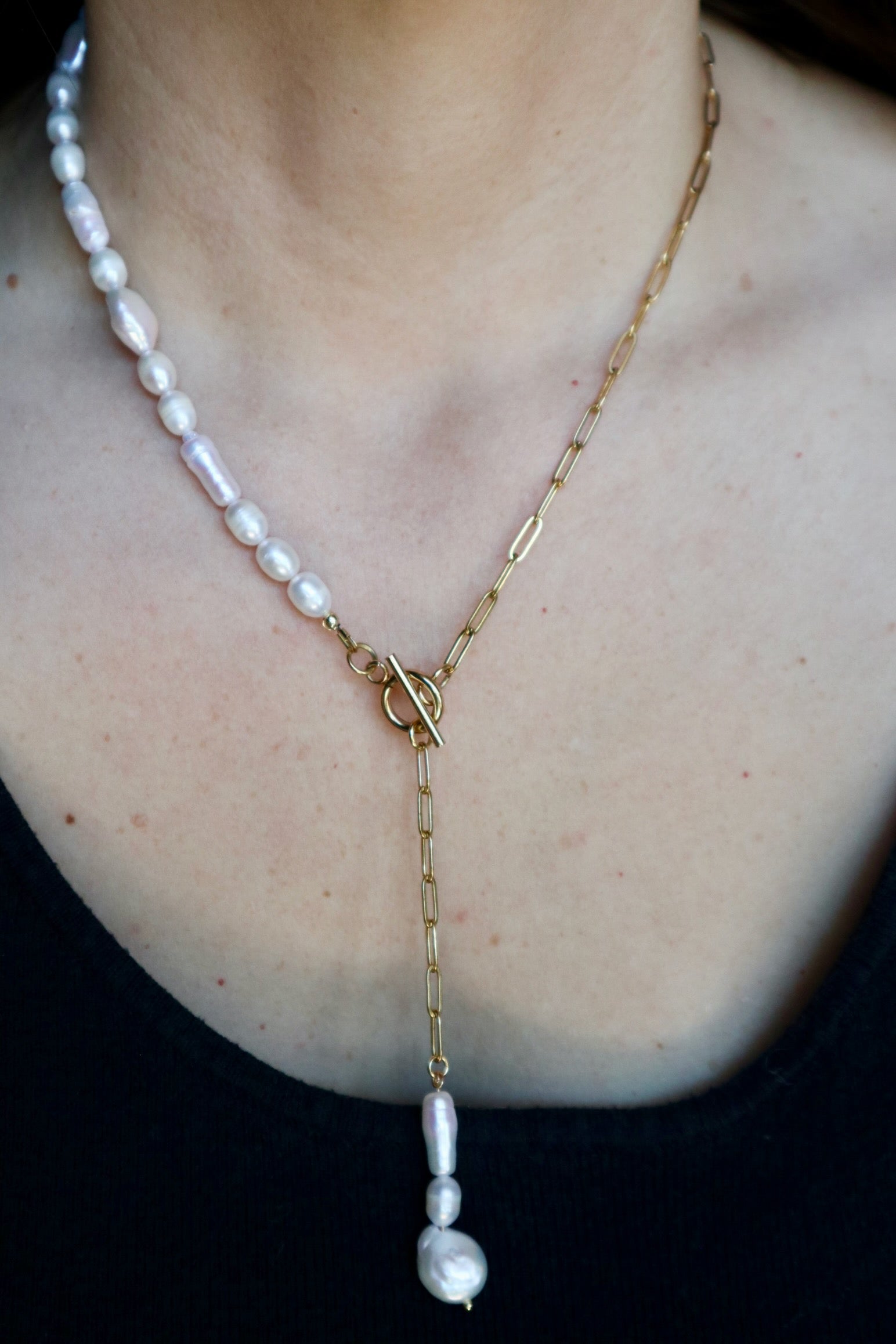 Auréad Designs Caelova lariat necklace worn on a black ribbed top, showing freshwater pearl strand on left, gold paperclip chain on right, toggle clasp at center front, and triple freshwater pearl pendant dropping below
