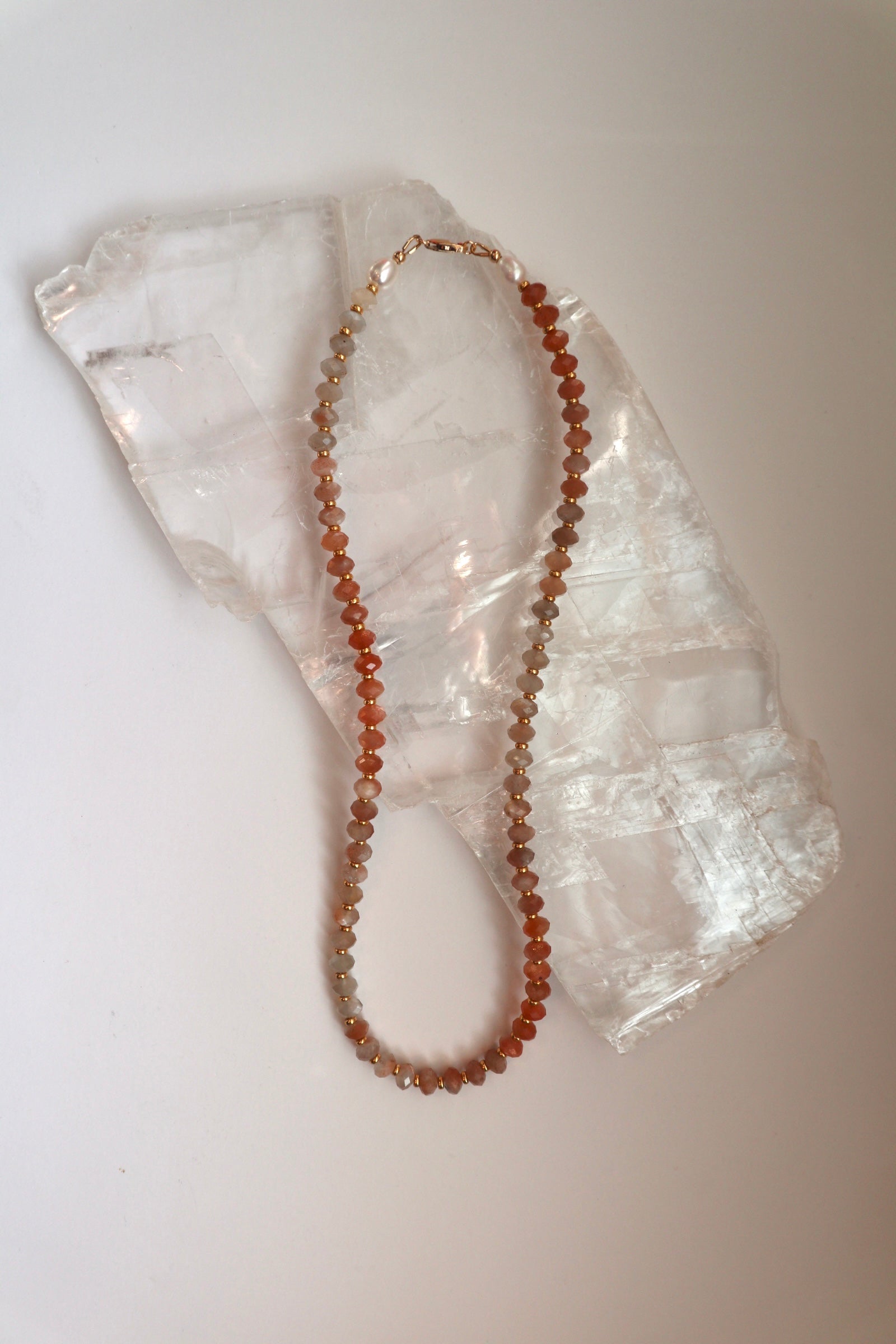 Calida necklace by Auréad Designs, featuring peach moonstone gemstone beads and freshwater pearls with galvanized gold spacer beads and a 14k gold-filled clasp, laid flat on a white selenite slab