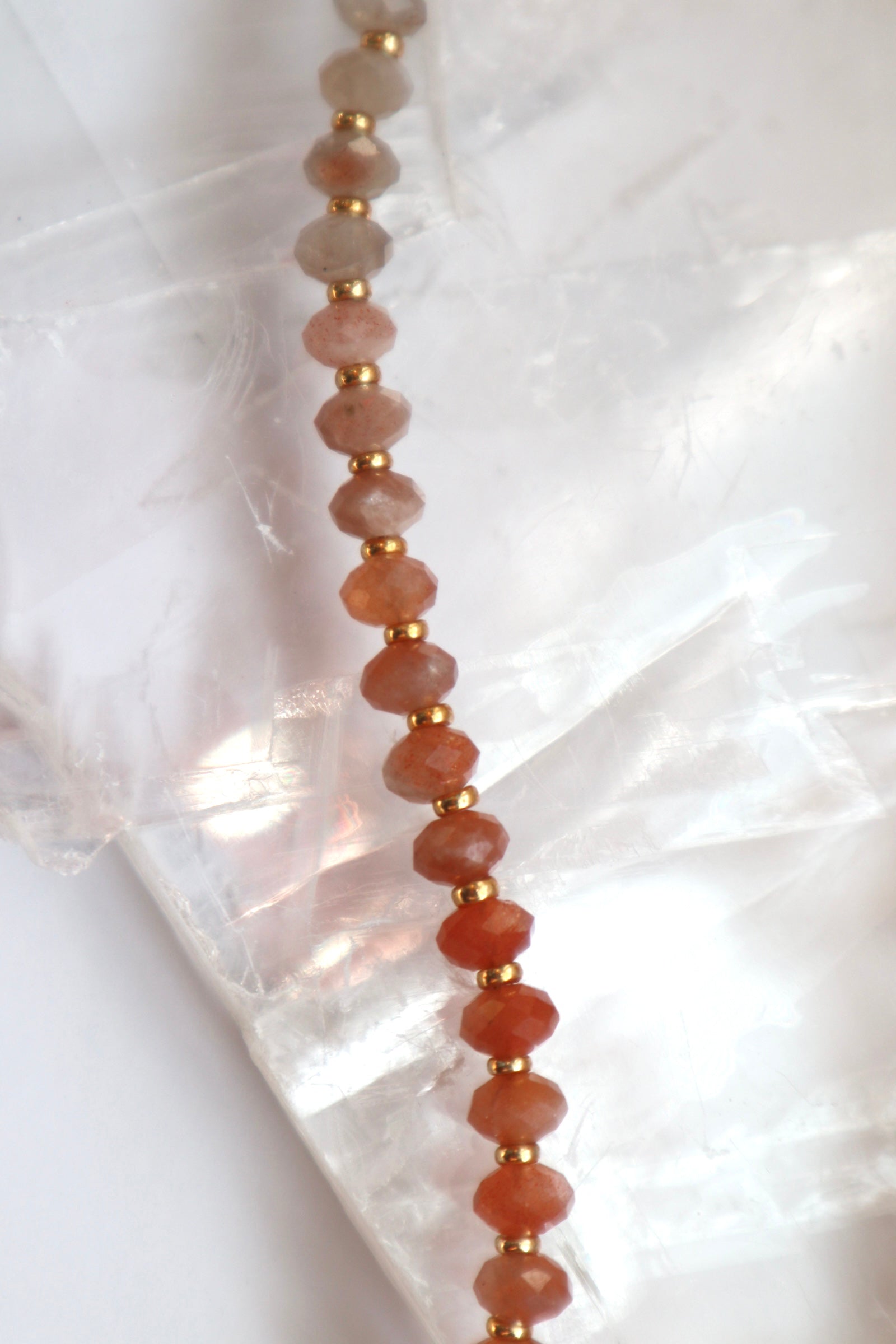 Close-up of the Calida necklace on a white selenite slab, showing the natural color gradient of peach moonstone beads from pale grey-peach to deeper coral tones with galvanized gold spacer beads
