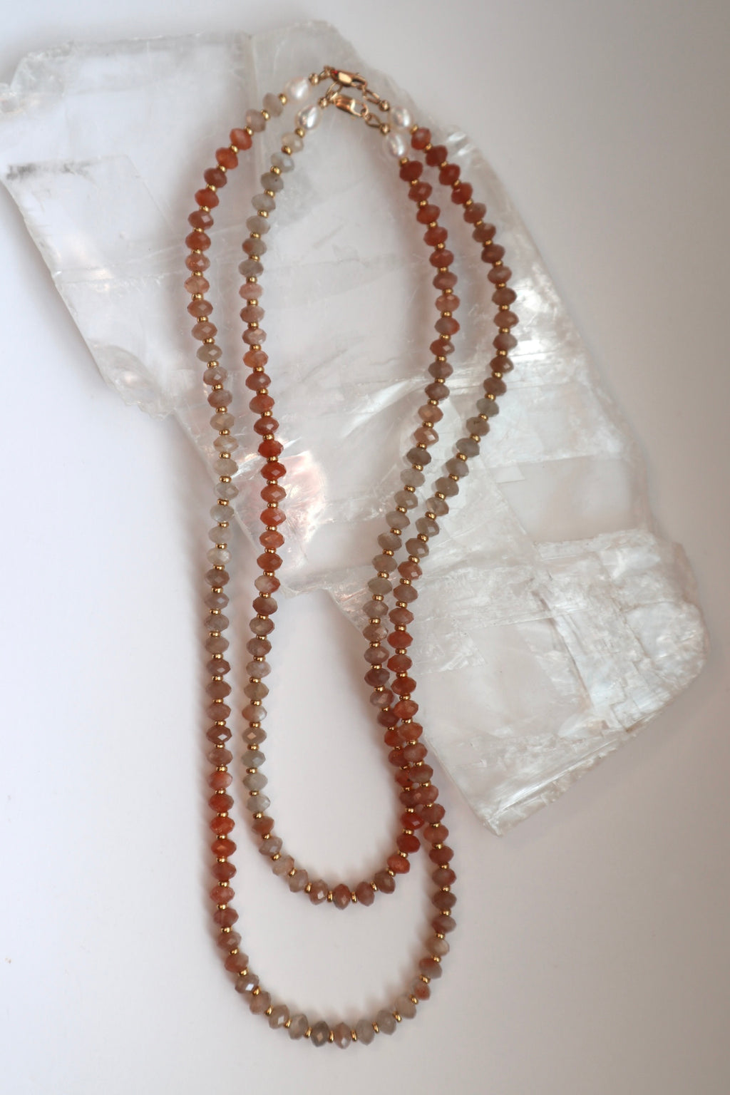 Calida necklace by Auréad Designs in both available lengths draped on a white selenite slab, showing the full color gradient of peach moonstone beads from pale grey-peach to deeper coral tones with gold spacer accents