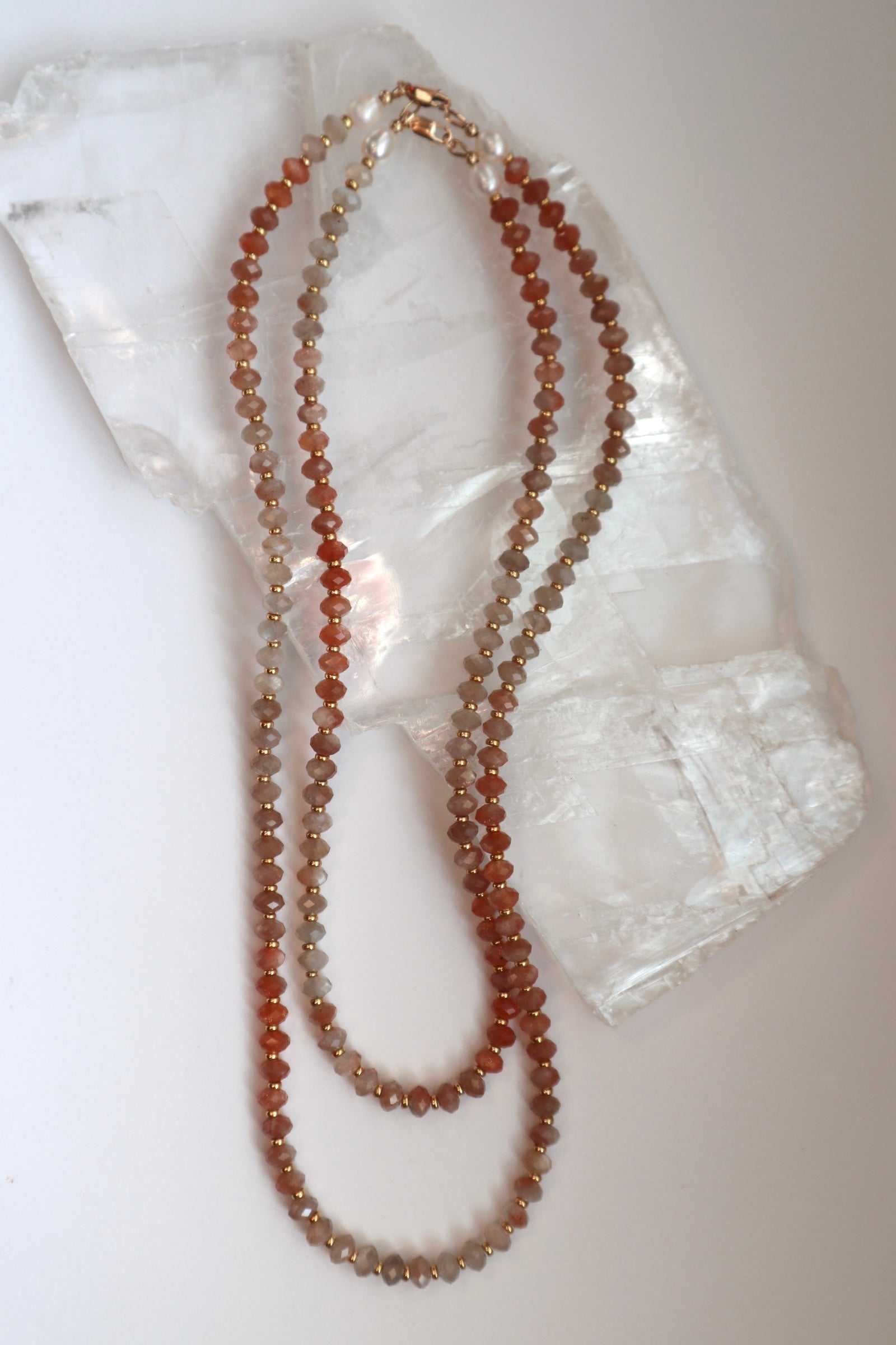 Calida necklace by Auréad Designs in both available lengths draped on a white selenite slab, showing the full color gradient of peach moonstone beads from pale grey-peach to deeper coral tones with gold spacer accents