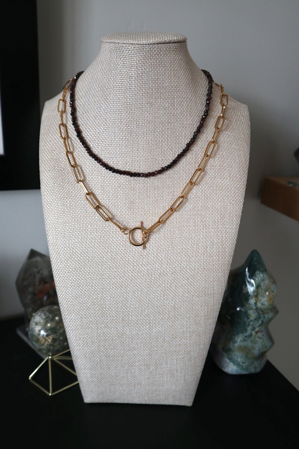 Catenne necklace by Auréad Designs displayed on a linen bust form, layered with a dark faceted gemstone necklace, with the gold toggle clasp as a focal point and crystal towers in the background