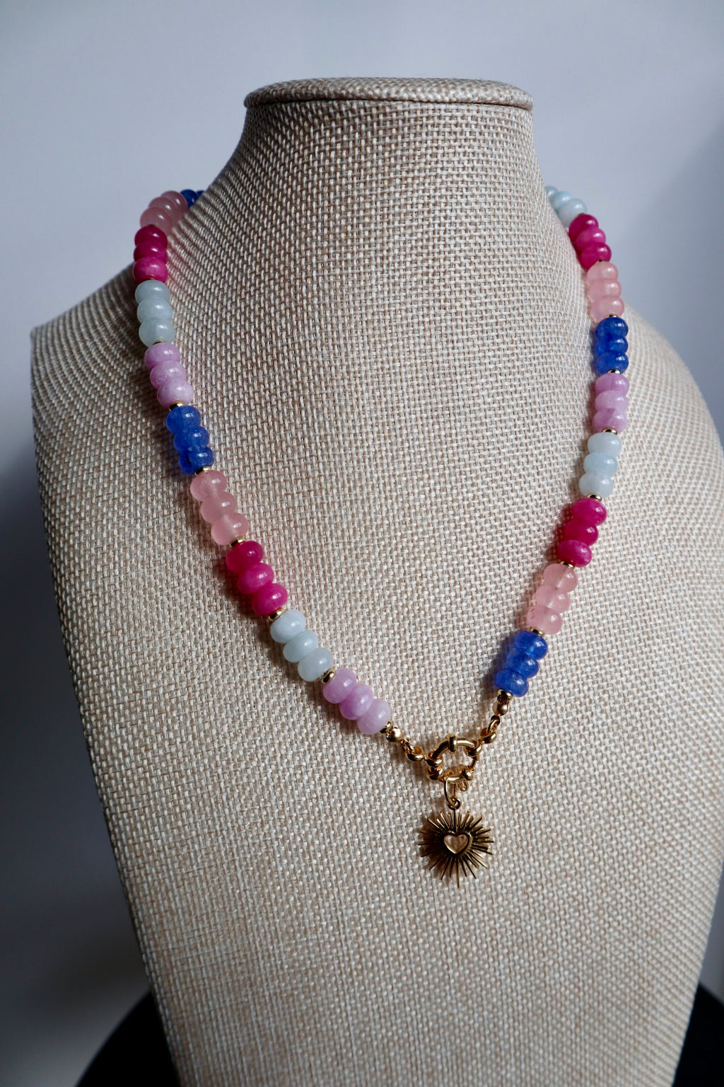 Cherise necklace by Auréad Designs on linen bust, showing the full strand of pink and purple jade rondelle beads with the gold radiant heart pendant as a focal point