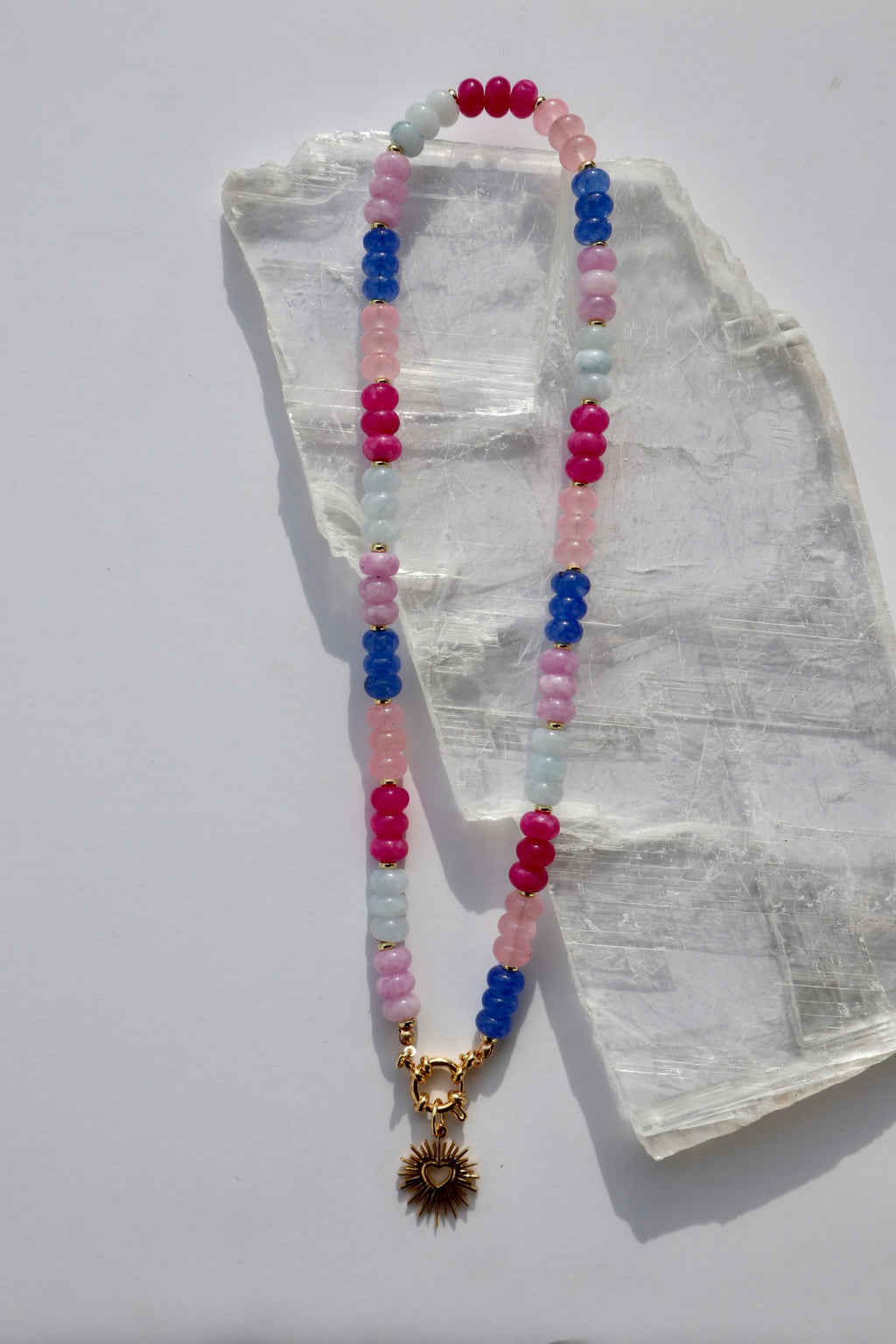 Cherise necklace by Auréad Designs, featuring dyed pink and purple jade rondelle beads and a gold stainless steel radiant heart pendant with a sailor spring clasp, laid flat on a white selenite slab