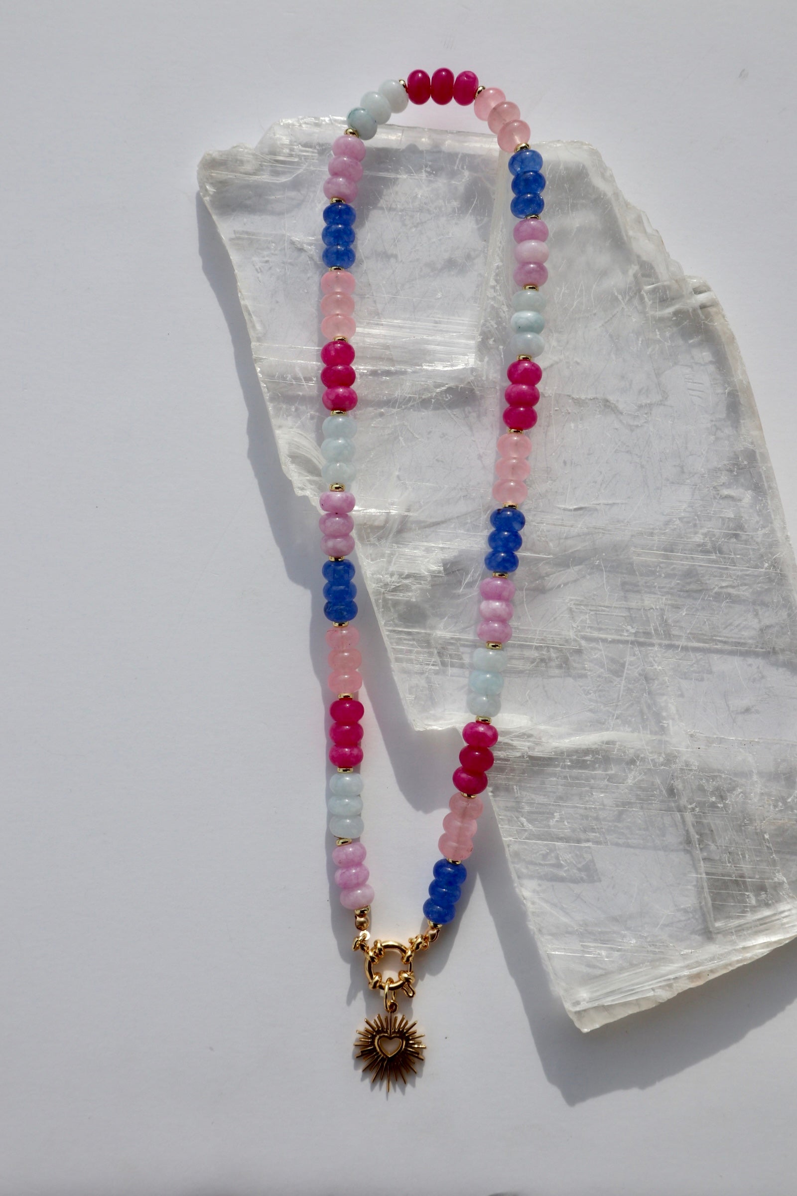 Cherise necklace by Auréad Designs, featuring dyed pink and purple jade rondelle beads and a gold stainless steel radiant heart pendant with a sailor spring clasp, laid flat on a white selenite slab