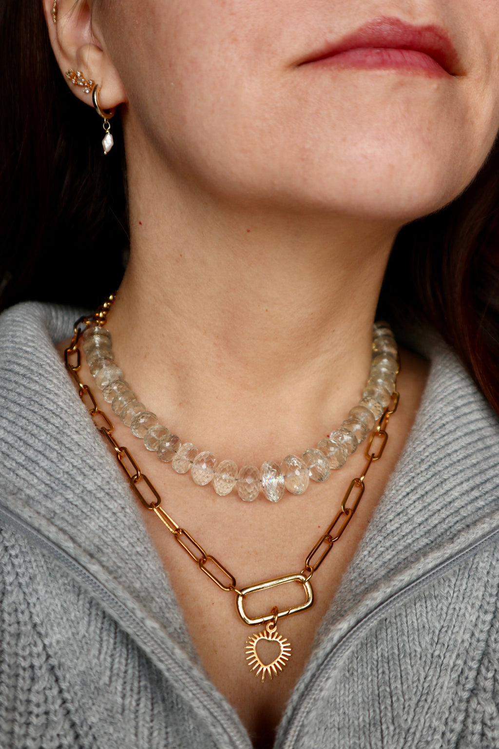 Clarae necklace by Auréad Designs layered on model with a gold paperclip chain and open sunburst heart charm pendant, styled with a gray knit to show a curated necklace stack