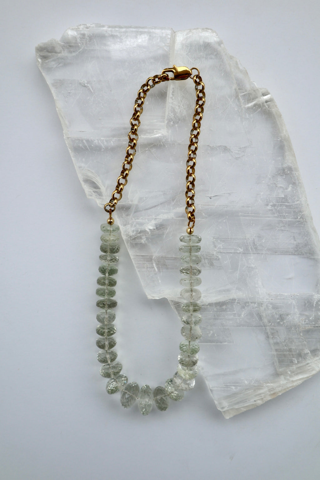 Clarae necklace by Auréad Designs, featuring graduated faceted green quartz gemstone beads transitioning to a gold stainless steel rolo chain with a lobster clasp, laid flat on a white selenite slab