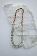 Clarae necklace by Auréad Designs, featuring graduated faceted green quartz gemstone beads transitioning to a gold stainless steel rolo chain with a lobster clasp, laid flat on a white selenite slab