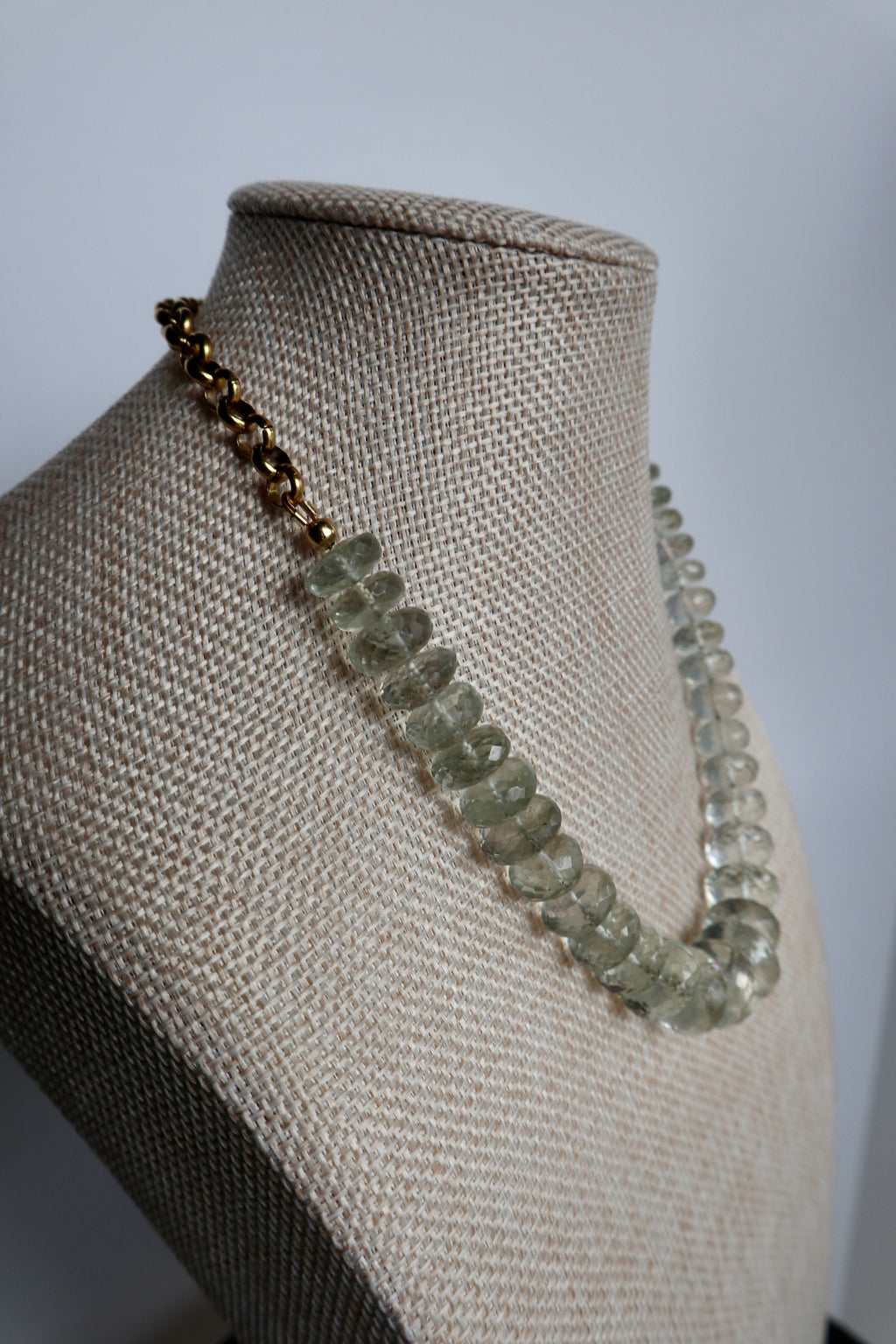 Clarae necklace by Auréad Designs displayed at an angle on a linen bust form, showing the full graduated faceted green quartz bead strand draping naturally with the gold stainless steel rolo chain at the shoulder
