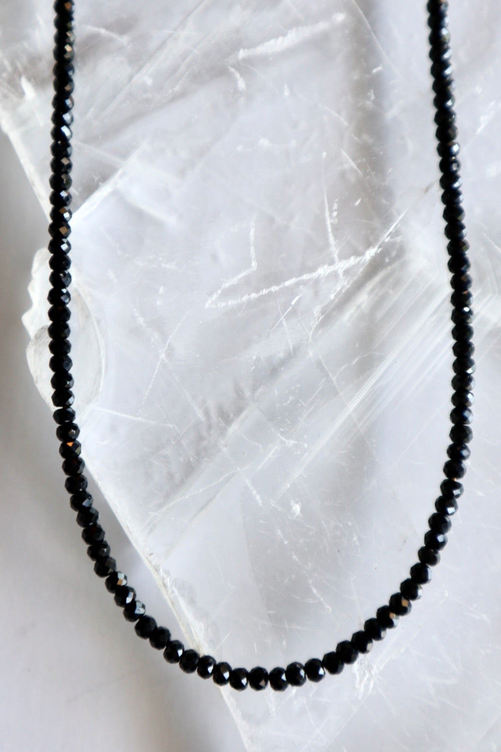 Auréad Designs Corvine necklace close-up of faceted rondelle black spinel beads on white selenite slab, showing micro-faceted surface and dark luminous finish