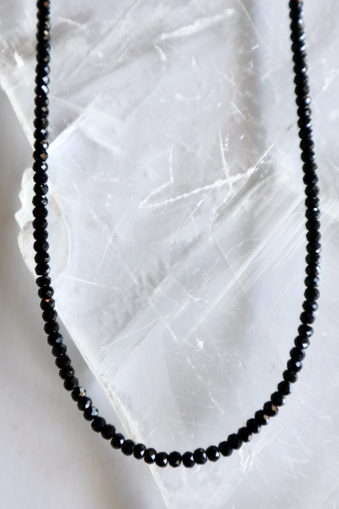 Auréad Designs Corvine necklace close-up of faceted rondelle black spinel beads on white selenite slab, showing micro-faceted surface and dark luminous finish
