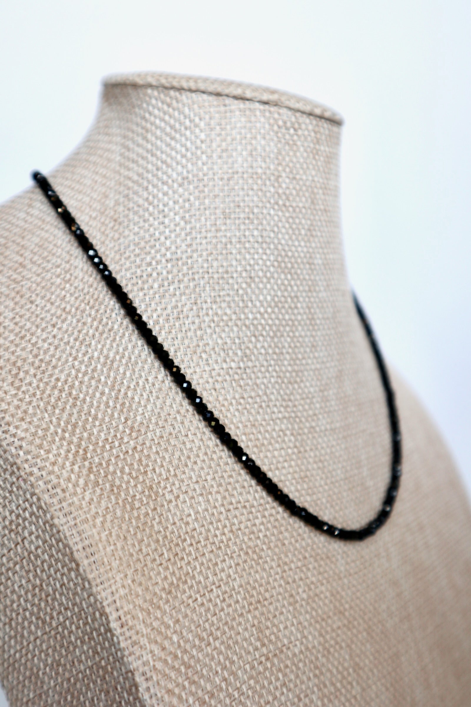 Auréad Designs Corvine necklace draped at an angle on linen bust form, showing full strand of faceted black spinel beads against neutral linen texture