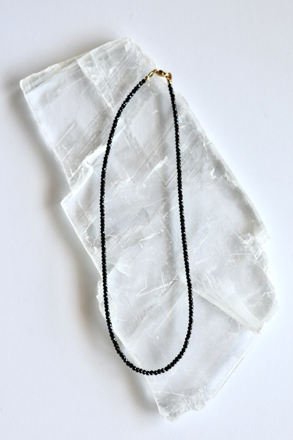 Auréad Designs Corvine necklace laid flat on white selenite slab, showing full strand of 3x2mm faceted rondelle black spinel beads with gold stainless steel lobster clasp