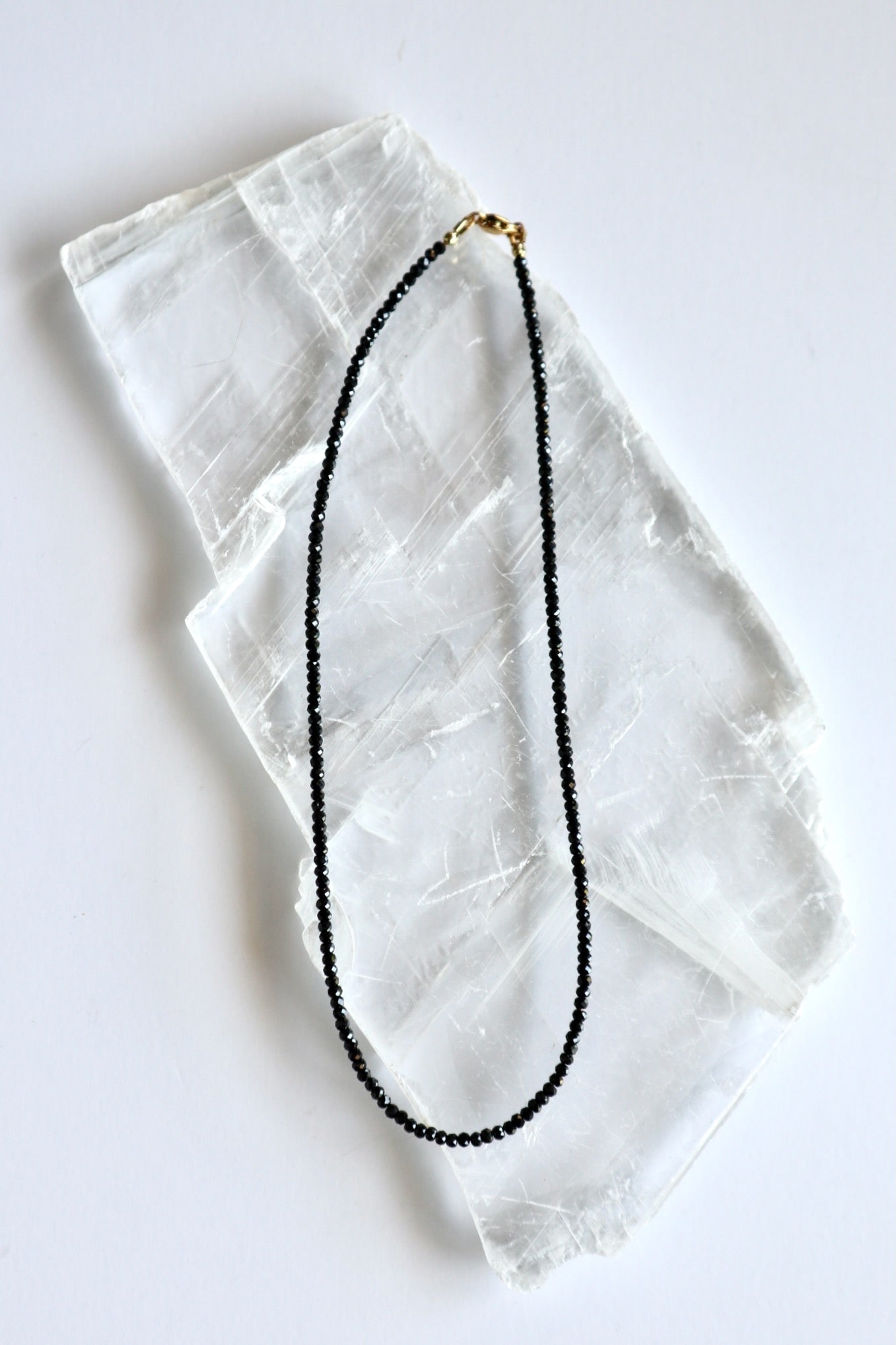 Auréad Designs Corvine necklace laid flat on white selenite slab, showing full strand of 3x2mm faceted rondelle black spinel beads with gold stainless steel lobster clasp