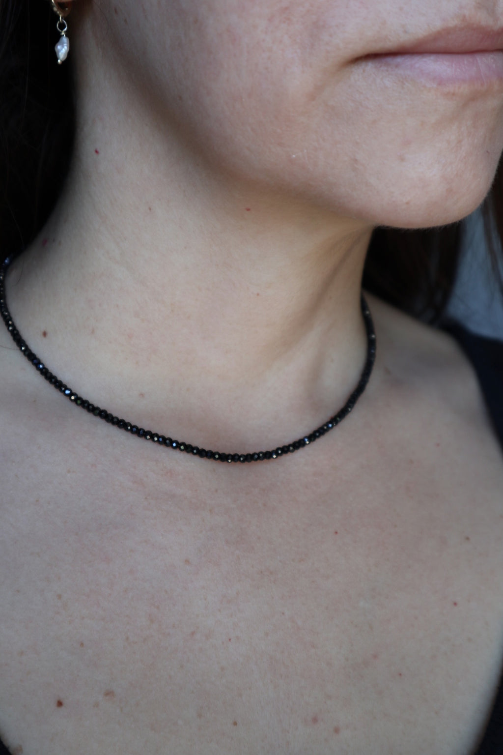 Auréad Designs Corvine necklace worn at collarbone length, showing faceted black spinel bead strand against bare skin with a pearl drop earring visible at left